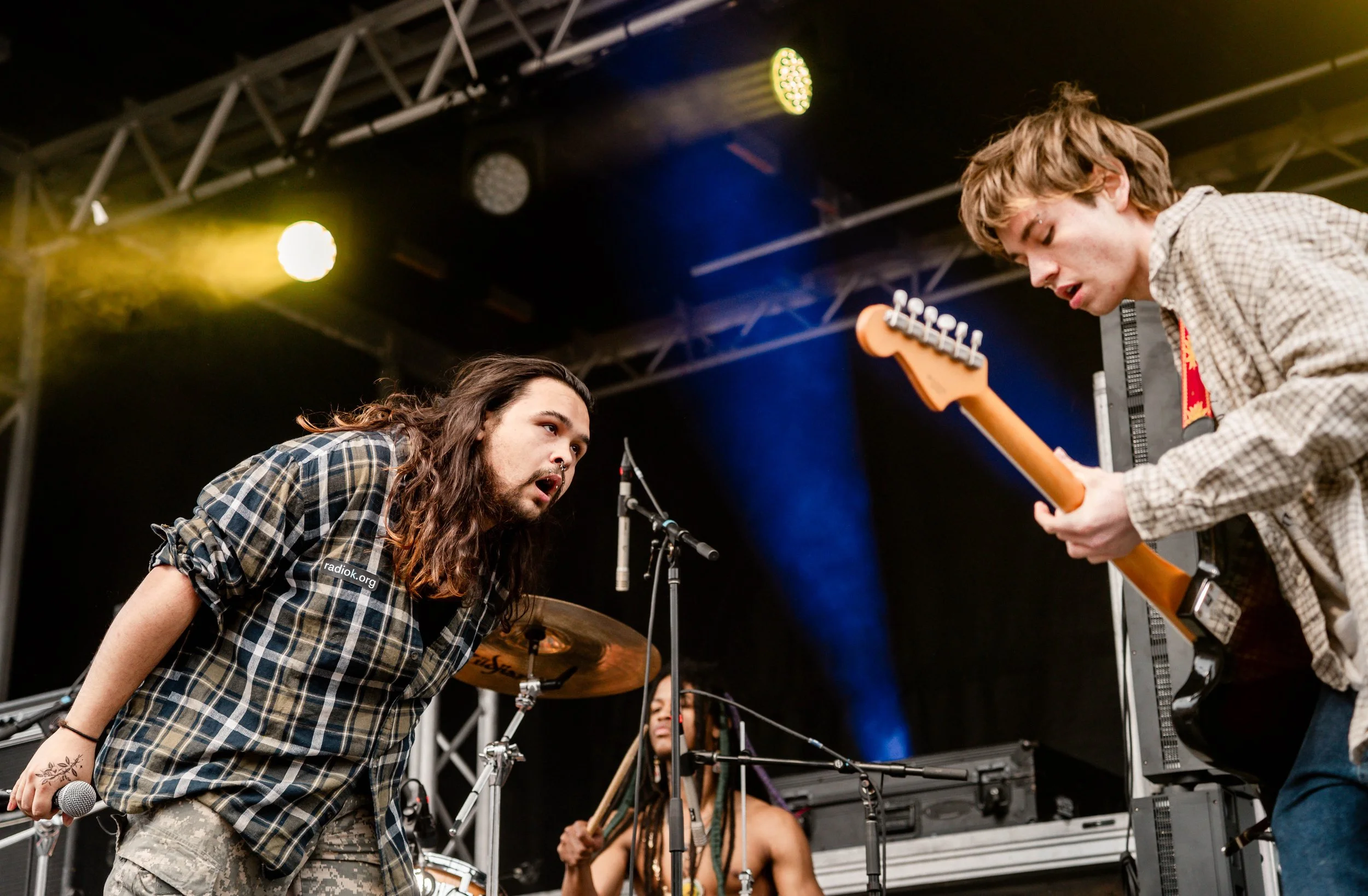 Two male musicians performing on stage, one with long hair wearing a plaid shirt singing into a microphone, the other with short hair playing an electric guitar, with a drummer in the background and stage lights overhead.