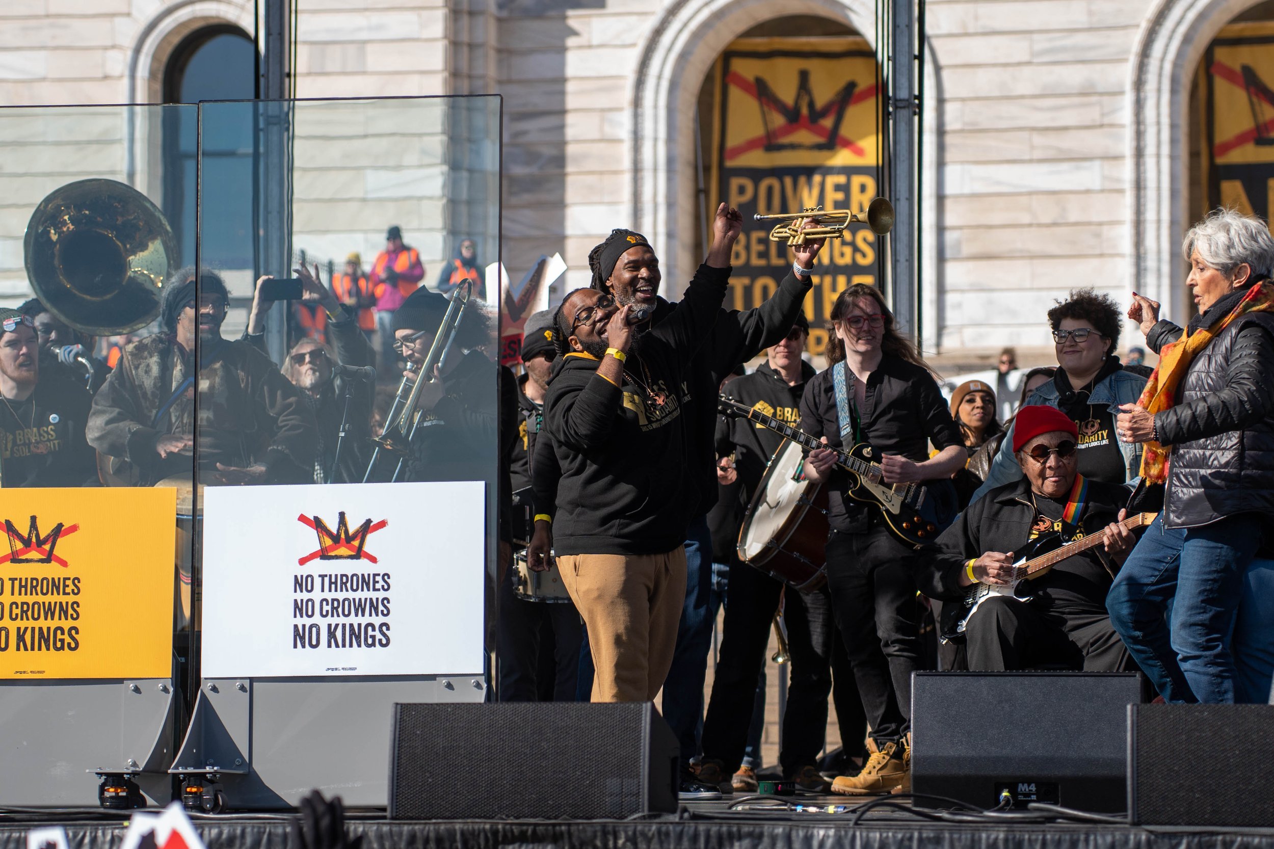A diverse group of people performing music and singing at a rally or protest outside a government building. The scene includes musicians with guitars, a person holding a trumpet, and signs reading 'No Thrones, No Crowns, No Kings.' The crowd appears engaged and animated, with some standing and speaking, against the backdrop of a historic building with large windows and banners.