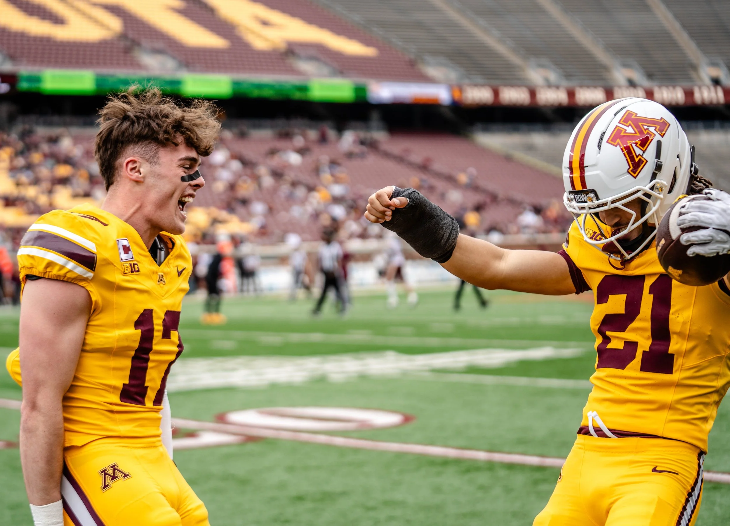Two football players wearing Minnesota Golden Gophers uniforms celebrating on the field. One player is smiling, and the other has a fist bump gesture.