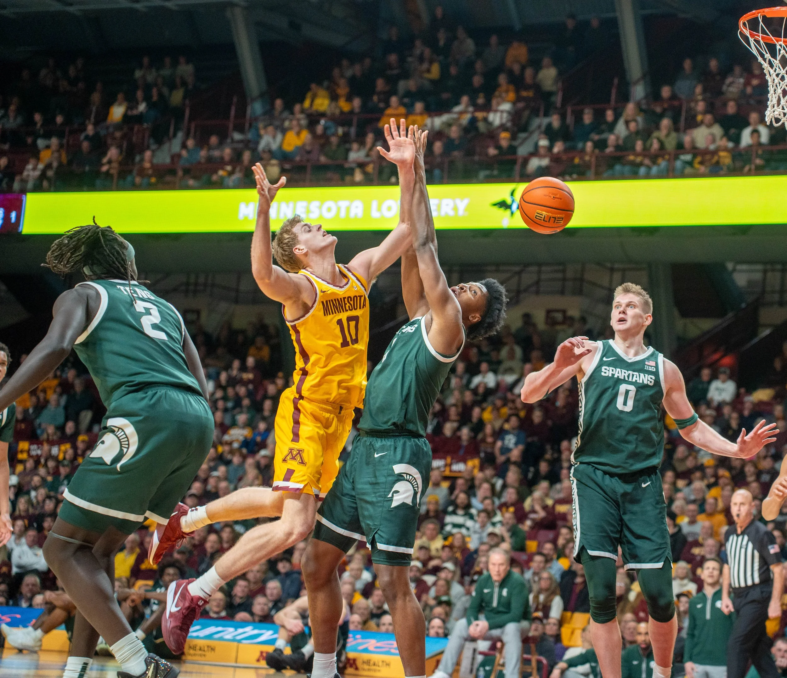 A basketball game with players jumping to contest a shot near the hoop, including players in yellow and green jerseys, with an audience in the background.