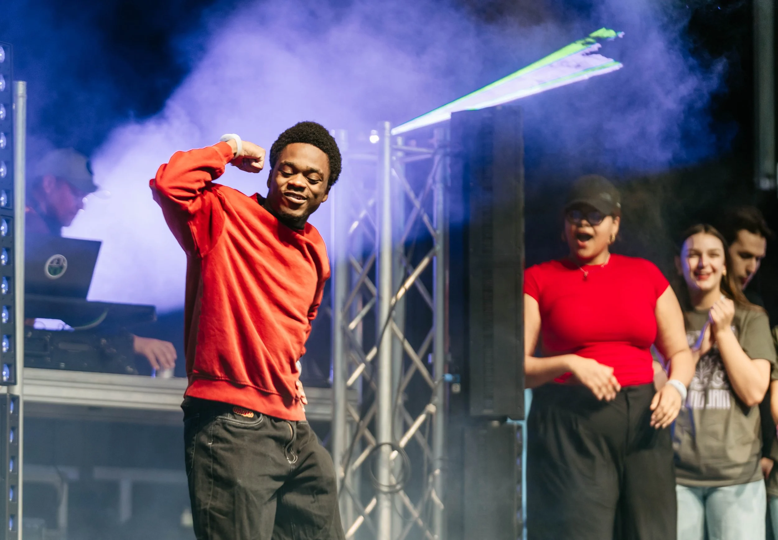 A young man in a red hoodie and black pants dancing on stage with a crowd watching, illuminated by stage lights and fog, at a lively event.