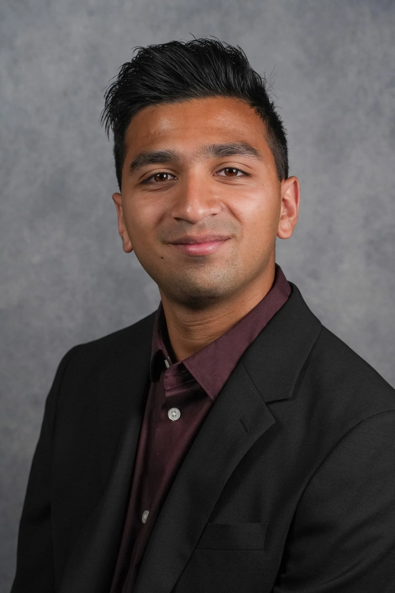 A professional headshot of a young man with dark hair, wearing a dark suit jacket and a maroon dress shirt, standing against a plain gray background.