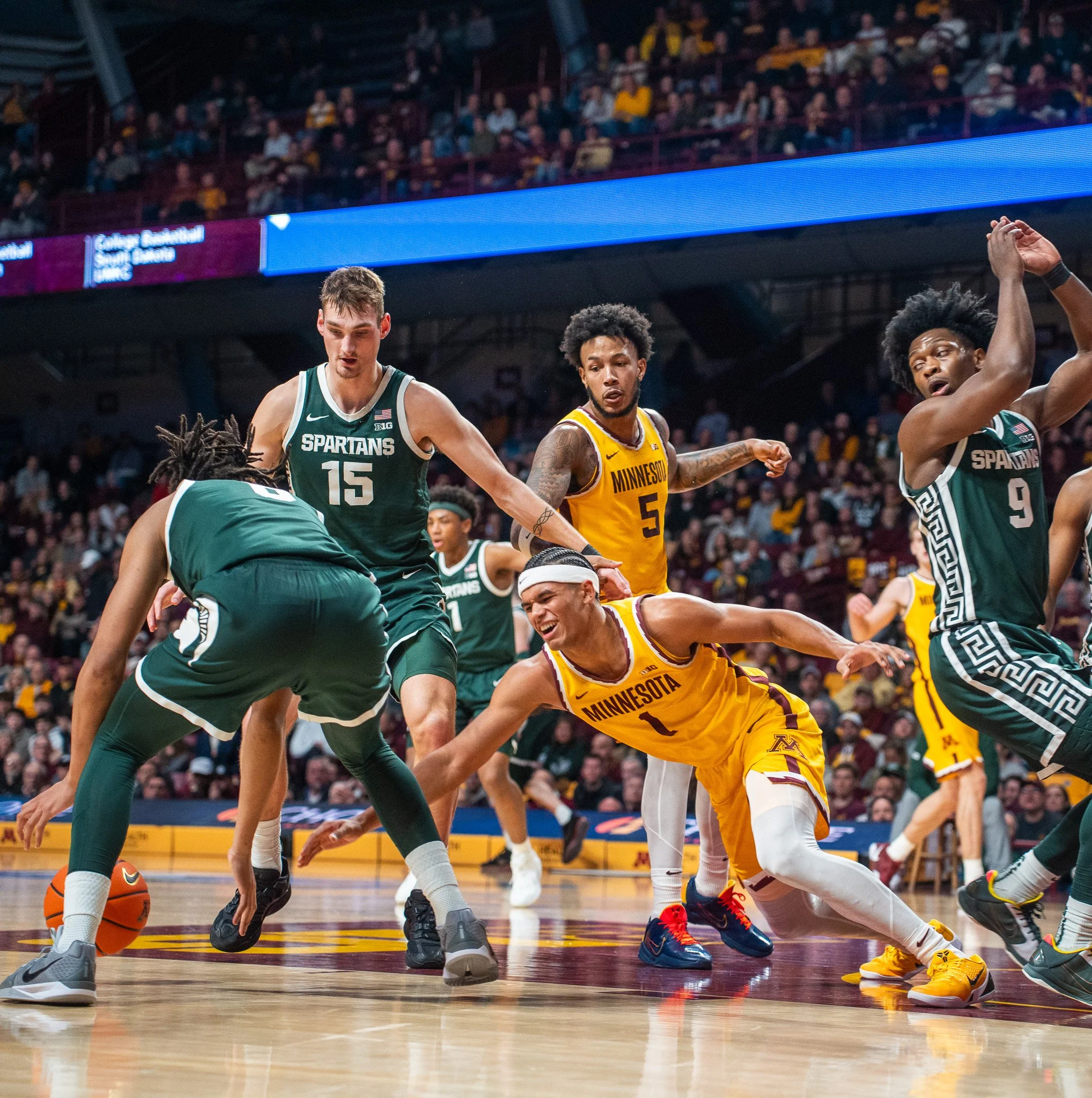 Basketball game with players from Michigan and Michigan State competing for possession of the ball on the court, with fans in the background.