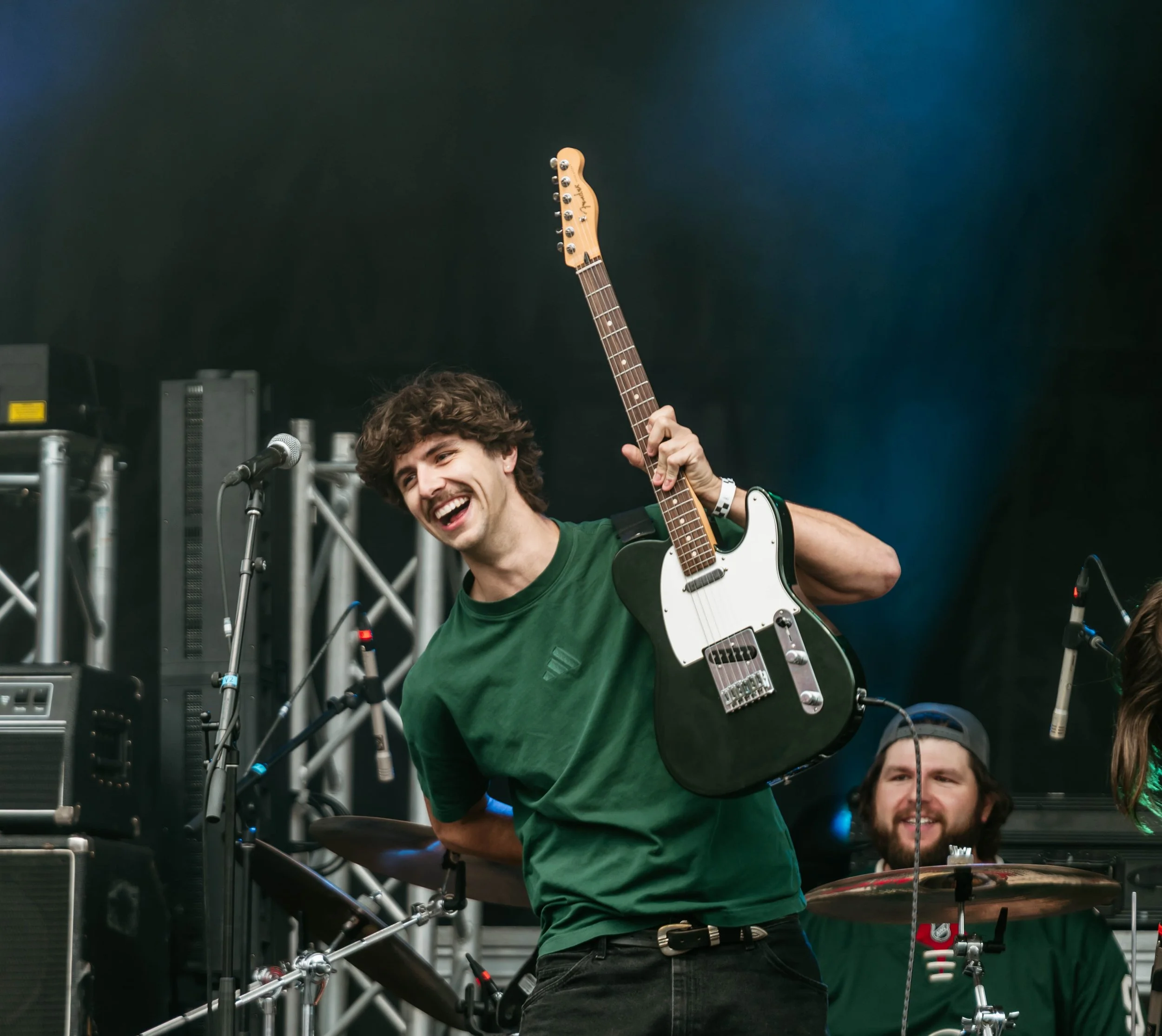 A young man with curly hair and a white watch is smiling and holding up an electric guitar, with a person partially visible sitting behind him, in an indoor setting with musical equipment.