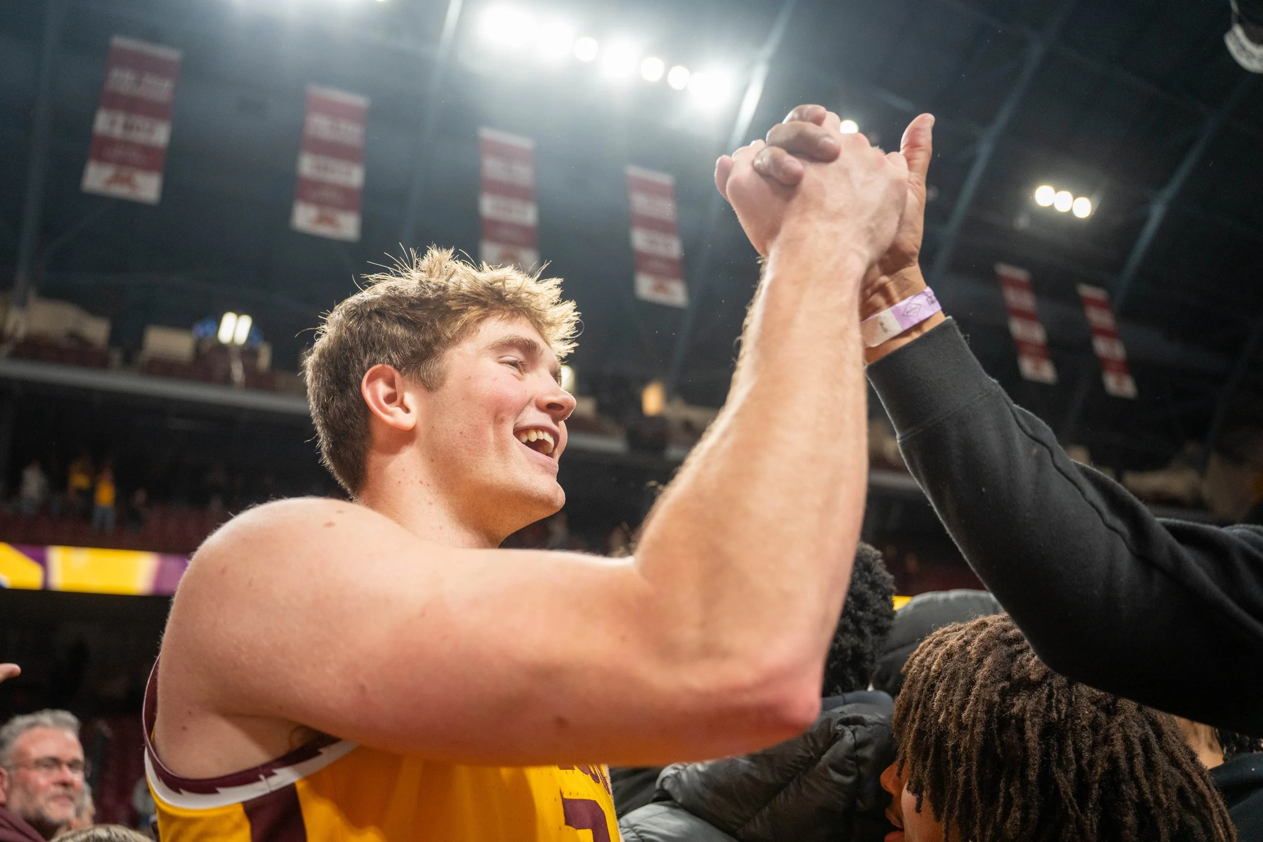A young male athlete celebrates with a handshake or high-five with a fan after a sporting event, smiling with closed eyes, in an indoor stadium with banners hanging from the ceiling.