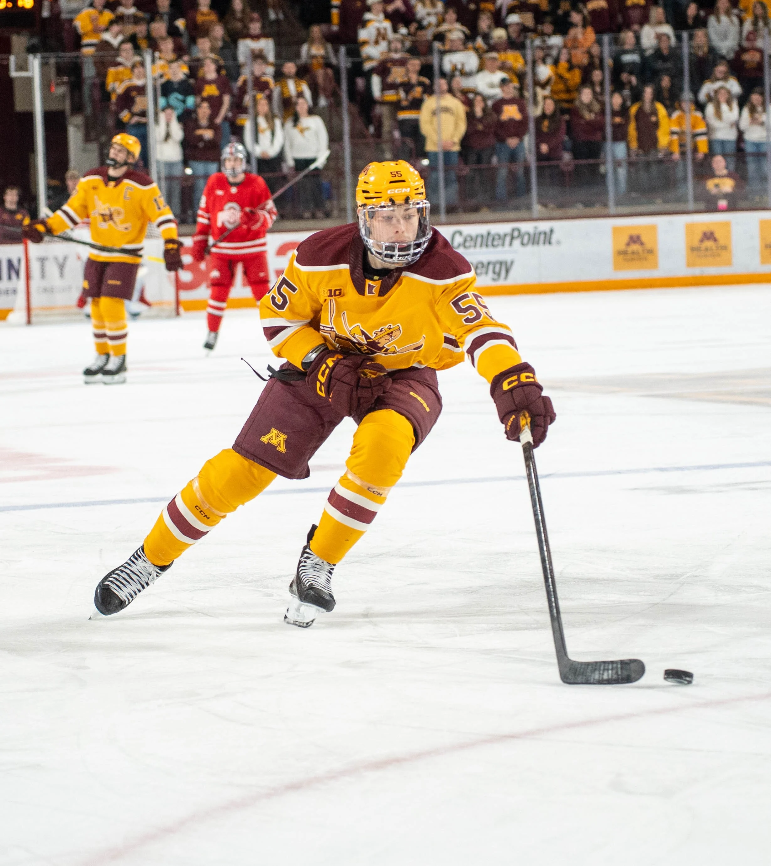An ice hockey player in yellow and maroon uniform with number 55 controlling a puck with a stick on the ice rink. Other players and a crowd of spectators are visible in the background.
