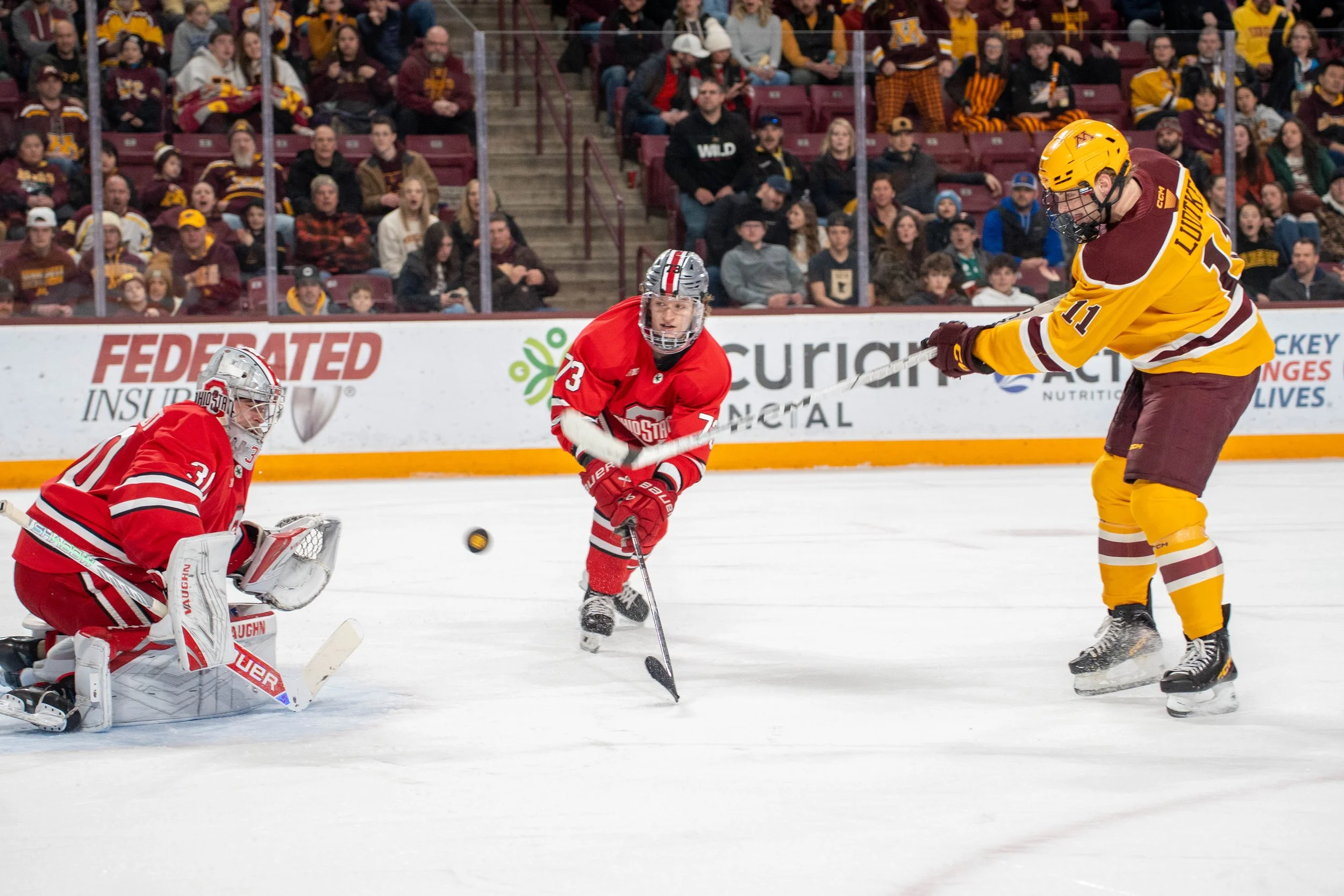 Hockey players in action during a game, with a goalie in red gear, a young player in red, and a player in a yellow jersey with the number 11, on ice rink with spectators watching from the stands.