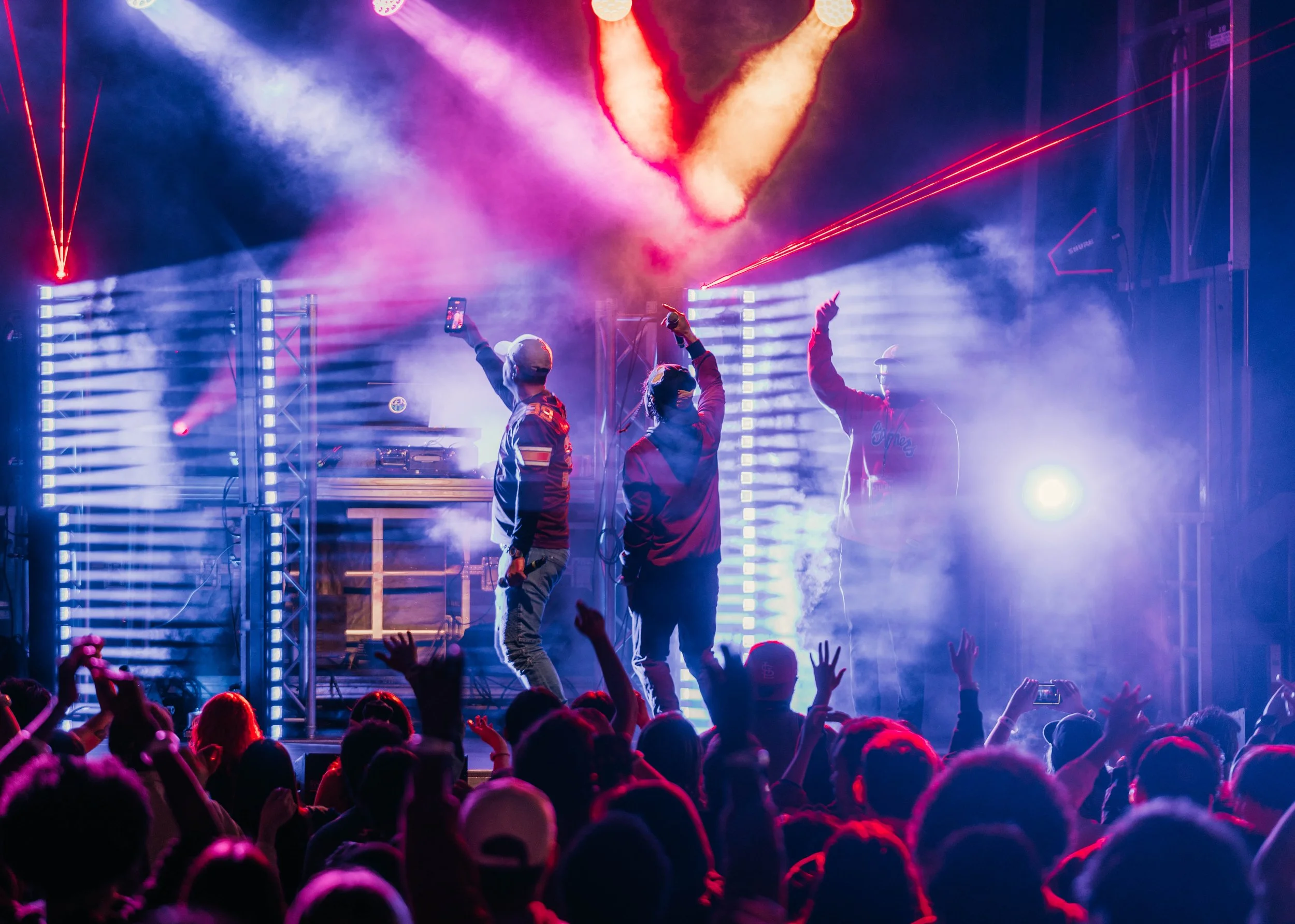 A live concert scene with performers on stage raising their hands, colorful lights, laser beams, and a crowd with hands raised.