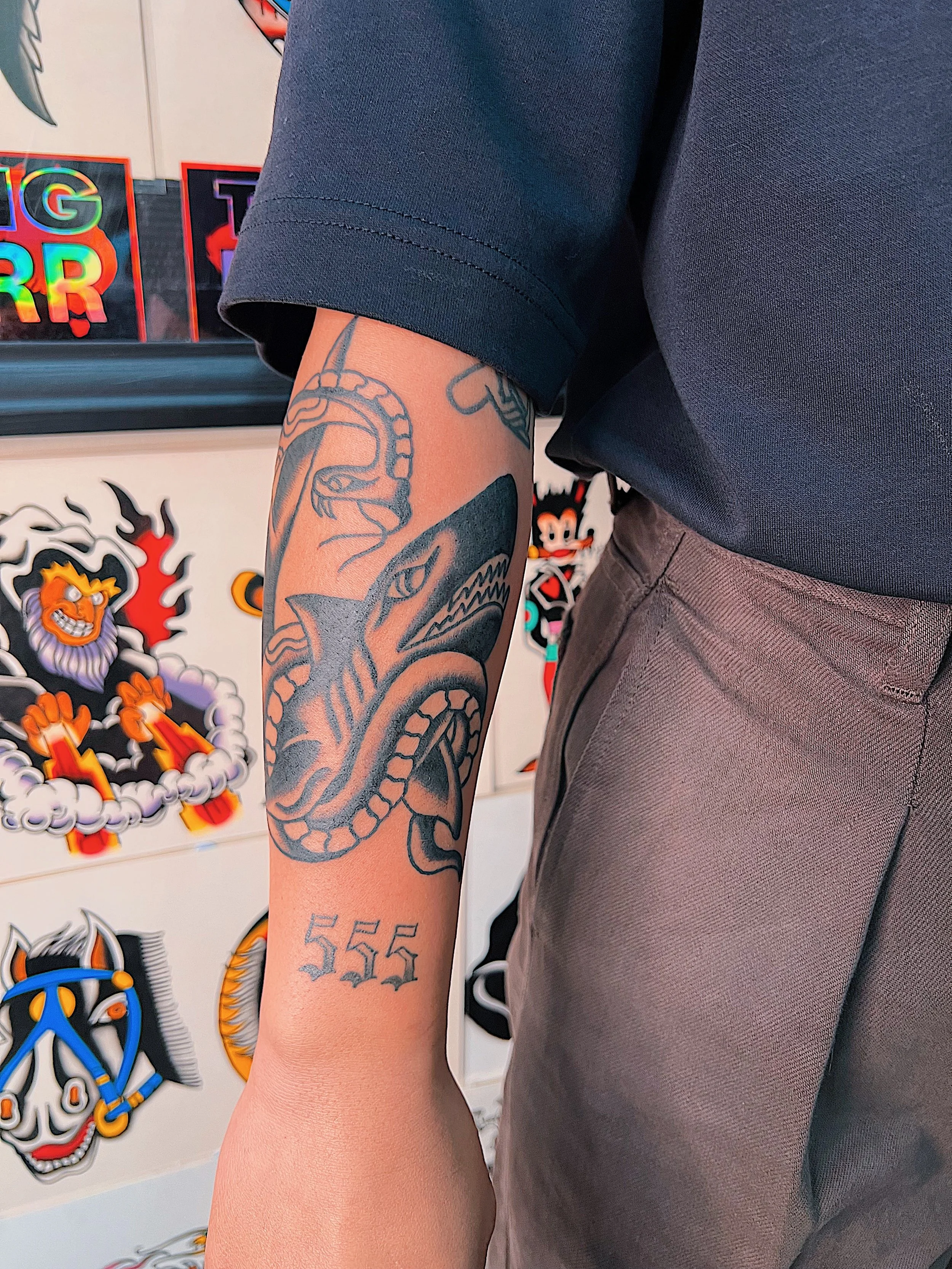 A person's arm with a tattoo of a snake and a shark, and the number 555 on their wrist, with various colorful tattoos visible. The background shows cartoon-style artwork on the wall.