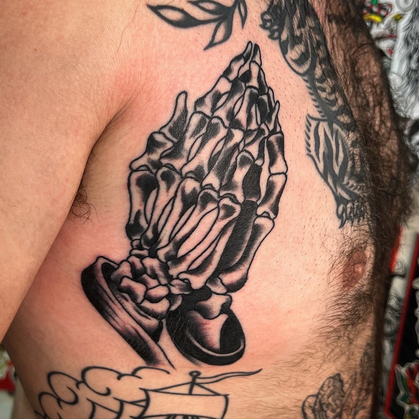 Tattoo of praying hands with skeletal fingers on man's chest surrounded by other tattoos of leaves and a ship.