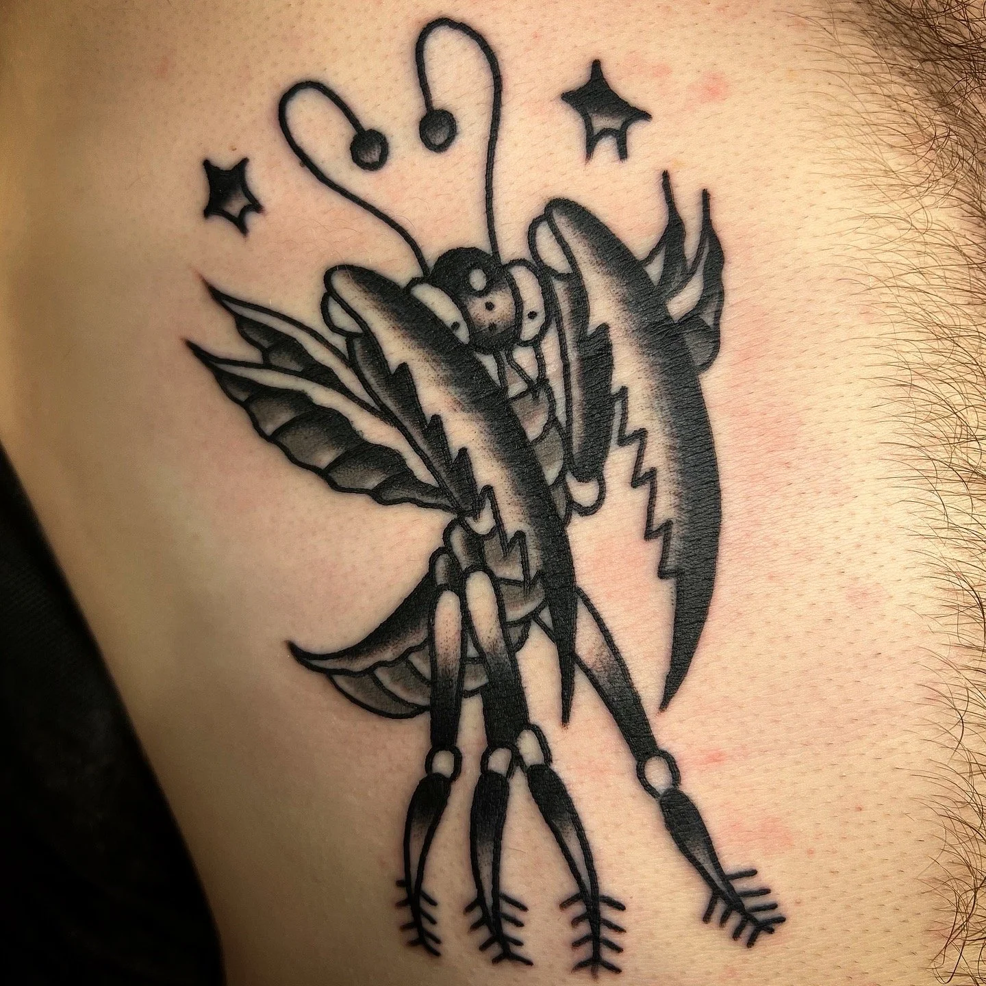 Black outline tattoo of a praying mantis with wings, antennae, and detailed legs, wearing headphones and surrounded by stars.