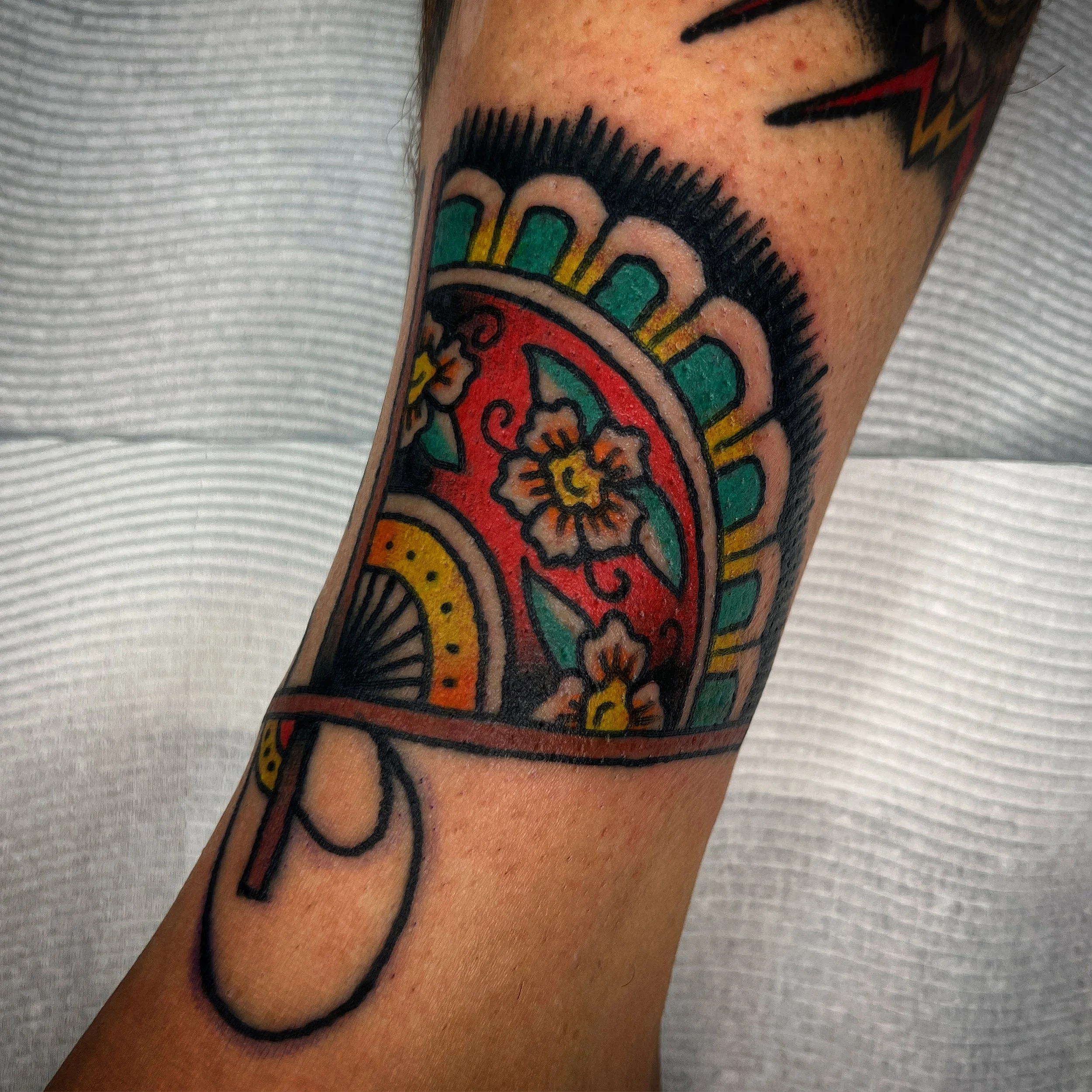Colorful tattoo of a decorated fan on a person's forearm.