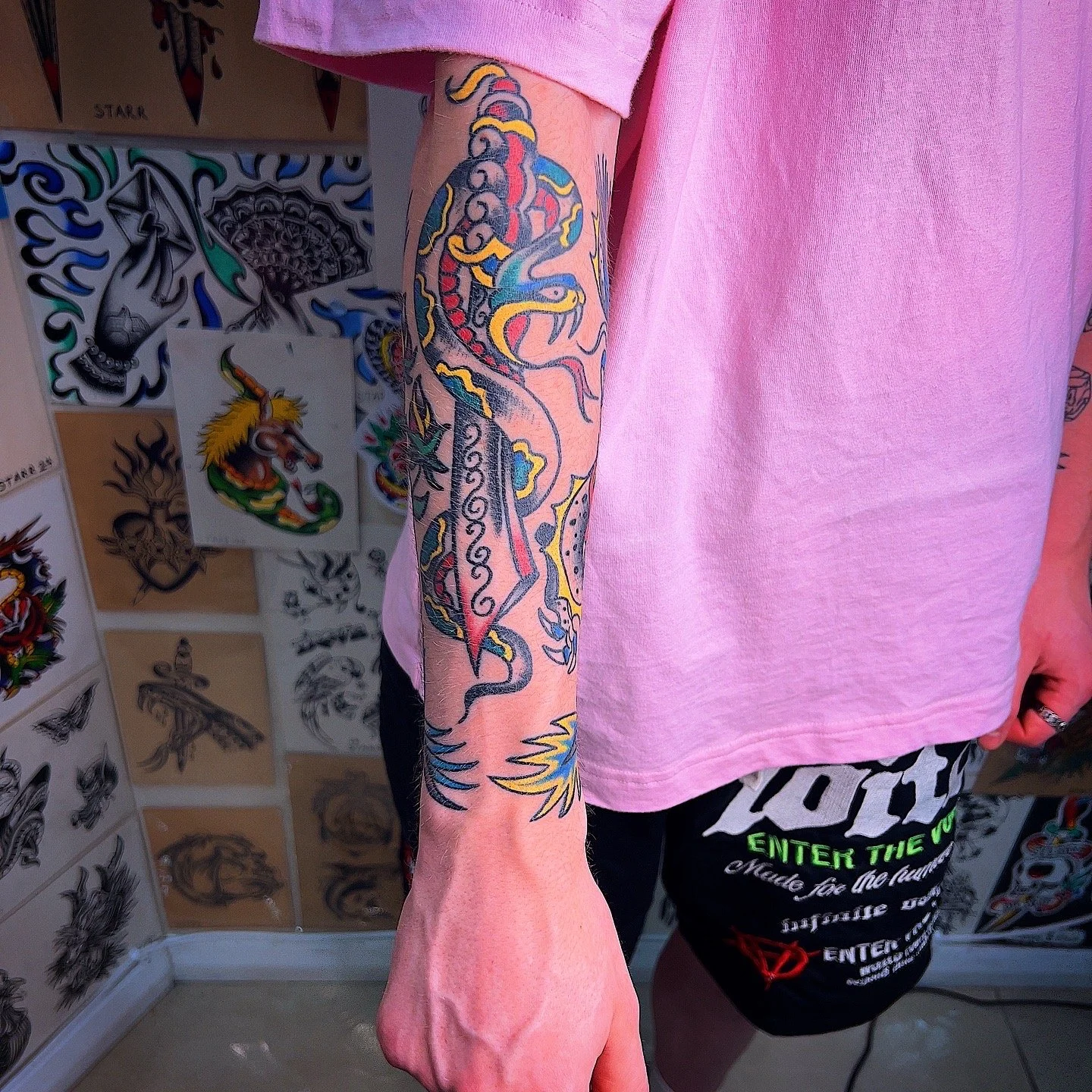 A person with colorful tattoos on their arm, featuring a dragon, standing in front of tattoo flash art on the wall.