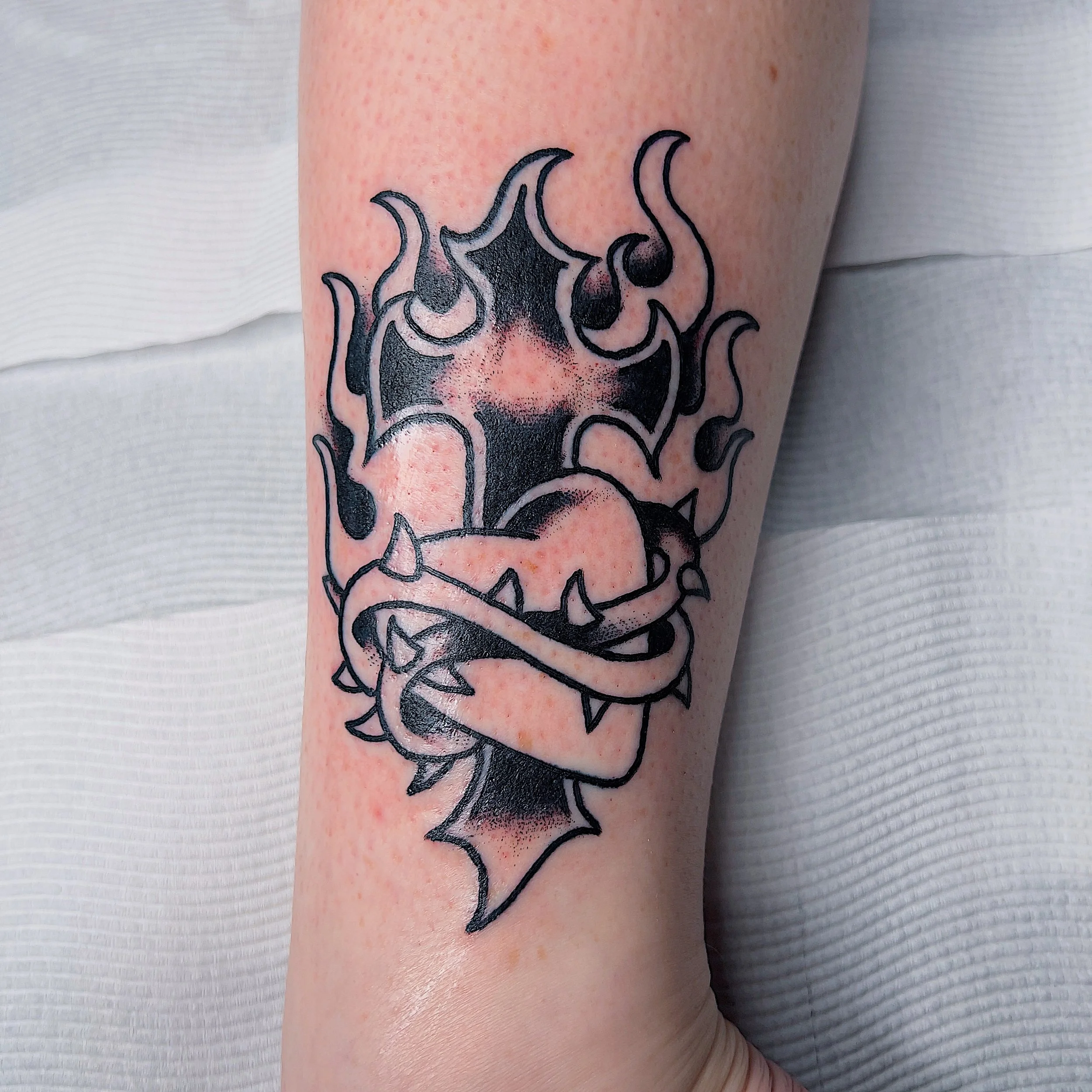 Black tribal-style tattoo of a cartoonish heart with flames and a band of spikes wrapped around it, on a person's forearm.