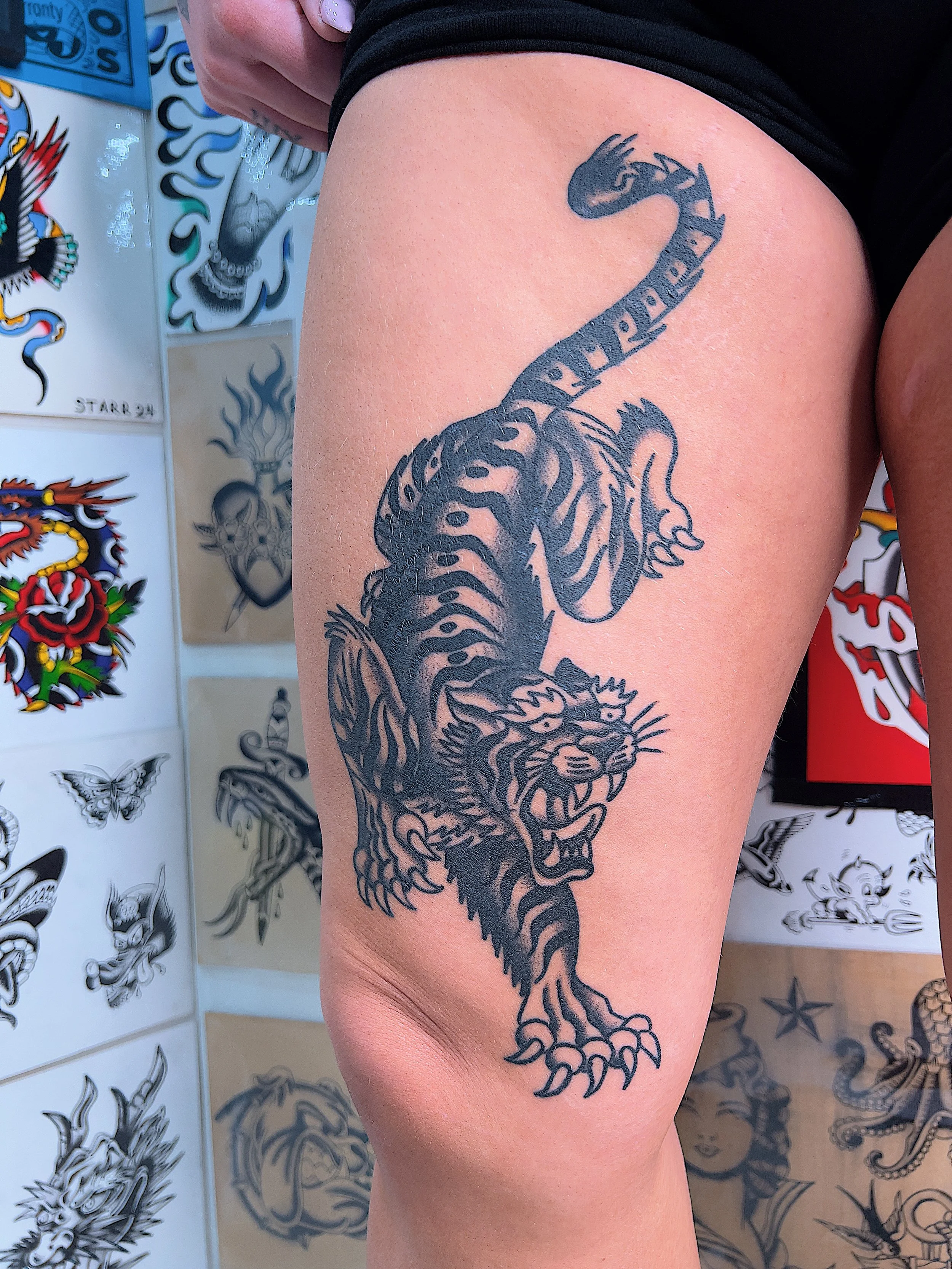 Black tattoo of a fierce tiger with stripes, showing its open mouth and teeth, on a person's thigh.