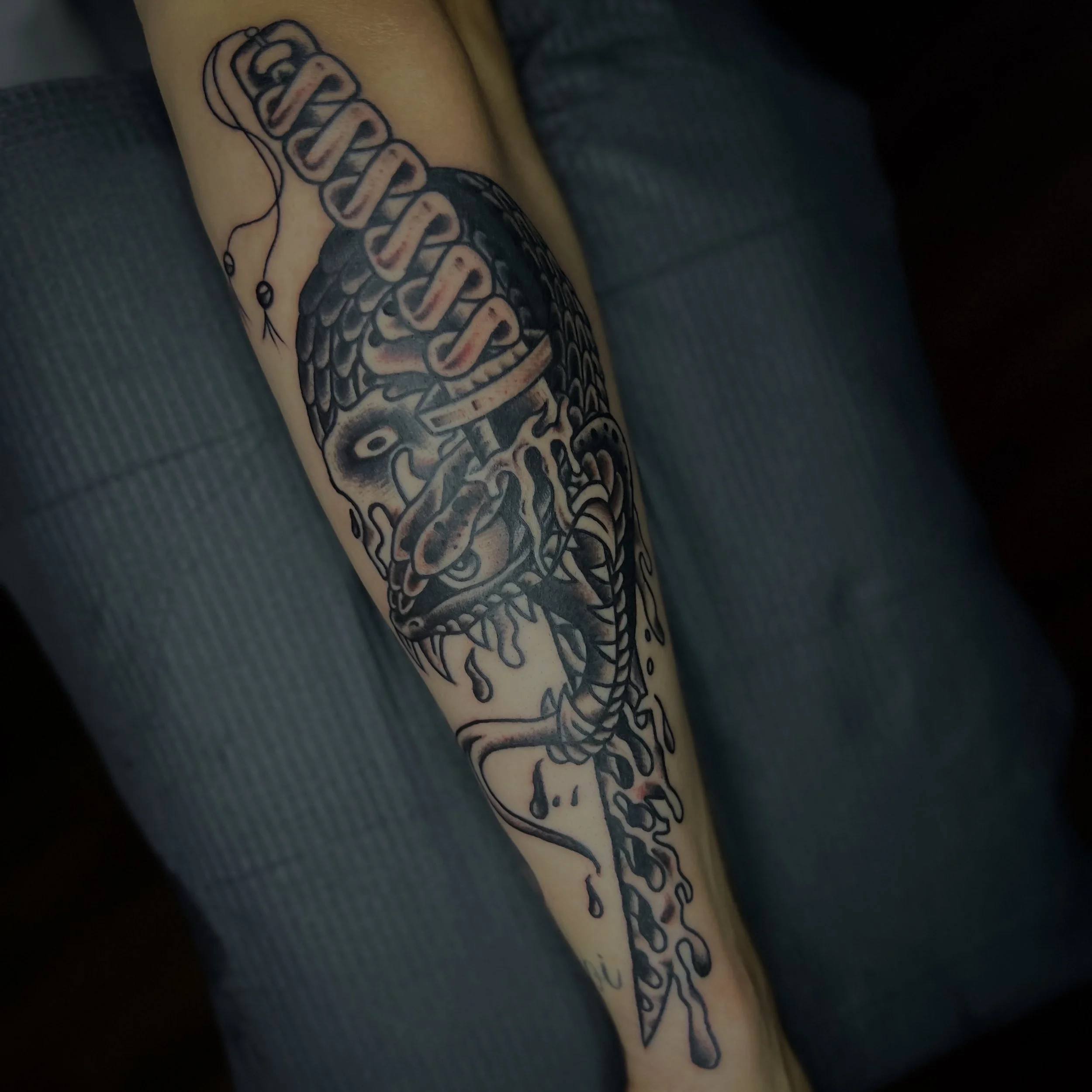 Tattoo of a snake entwined with a dagger on a person's forearm.