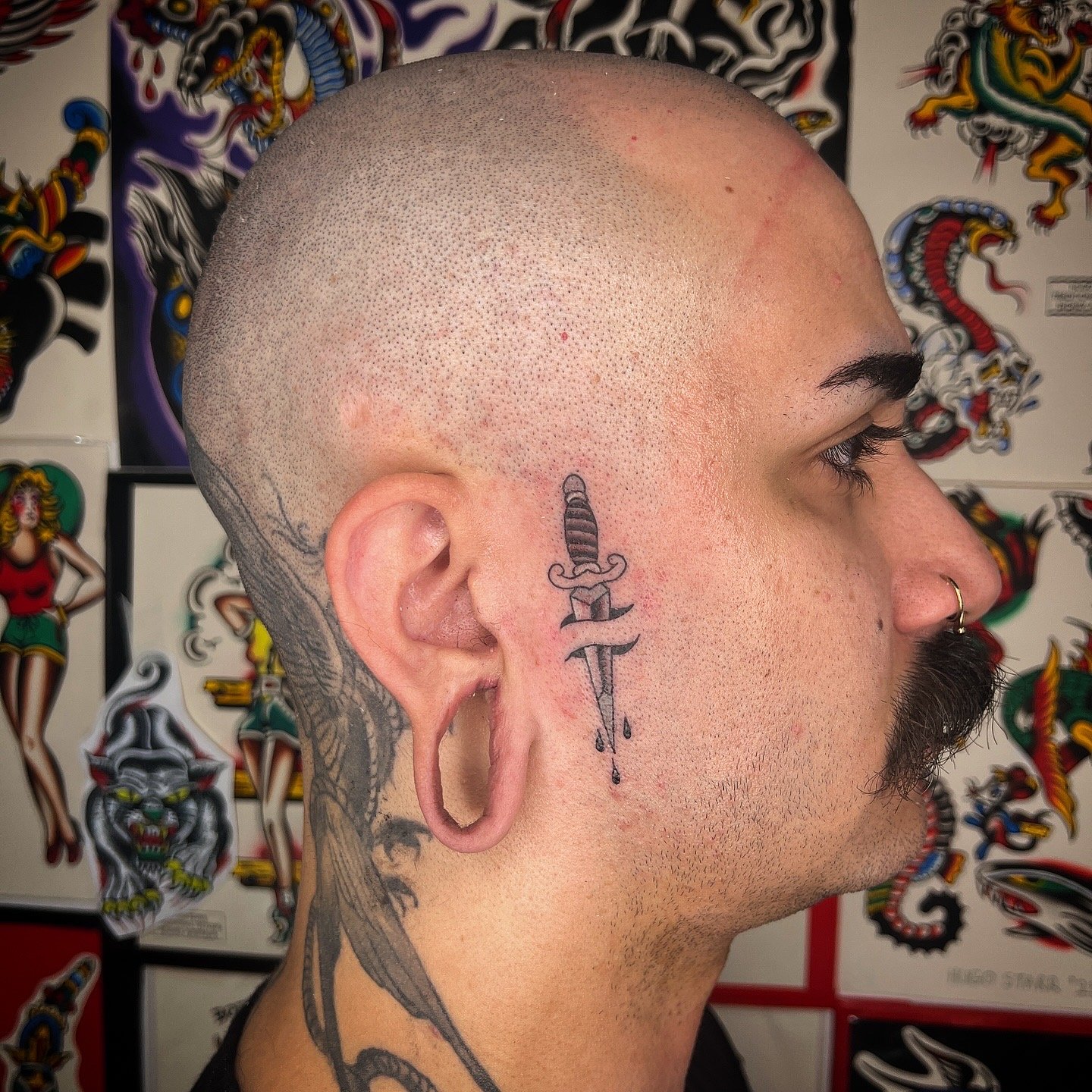 A close-up side profile of a man with a shaved head, facial hair, and tattoos, including a tattoo of a sword with a dripping effect behind the ear.