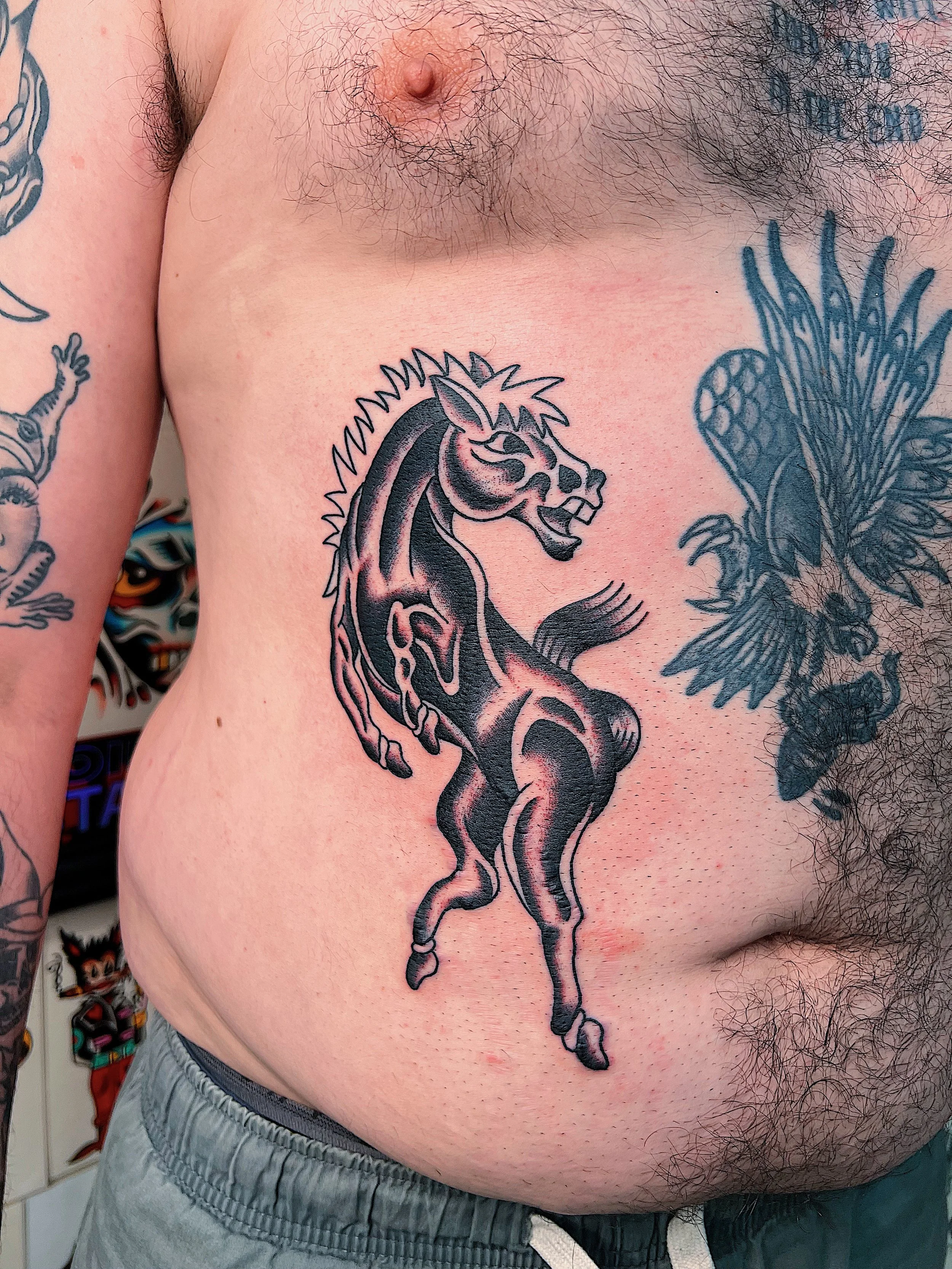 Tattoo of a black and gray stylized horse with a mane, tail, and muscular body on person's chest.