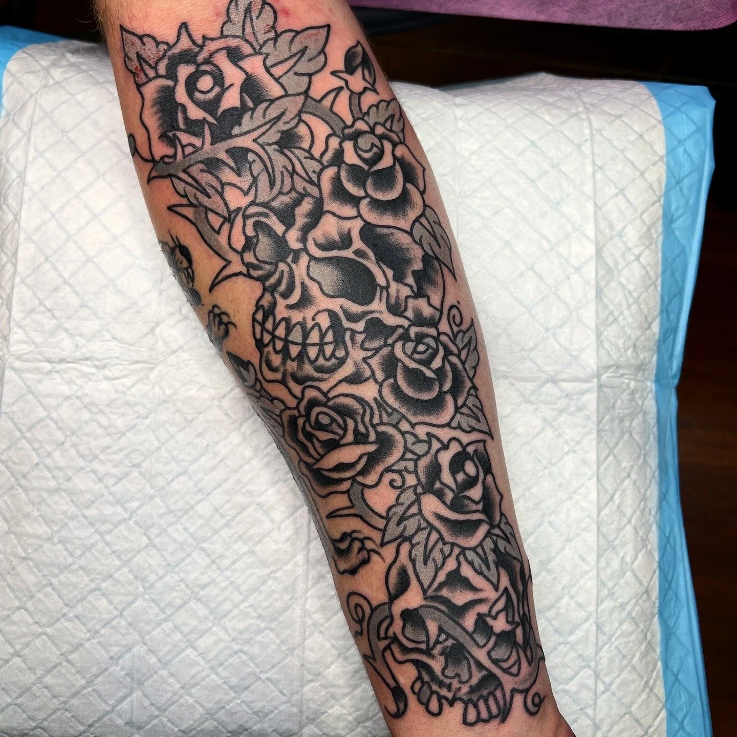 Tattoo of a skull surrounded by roses on a person's forearm.