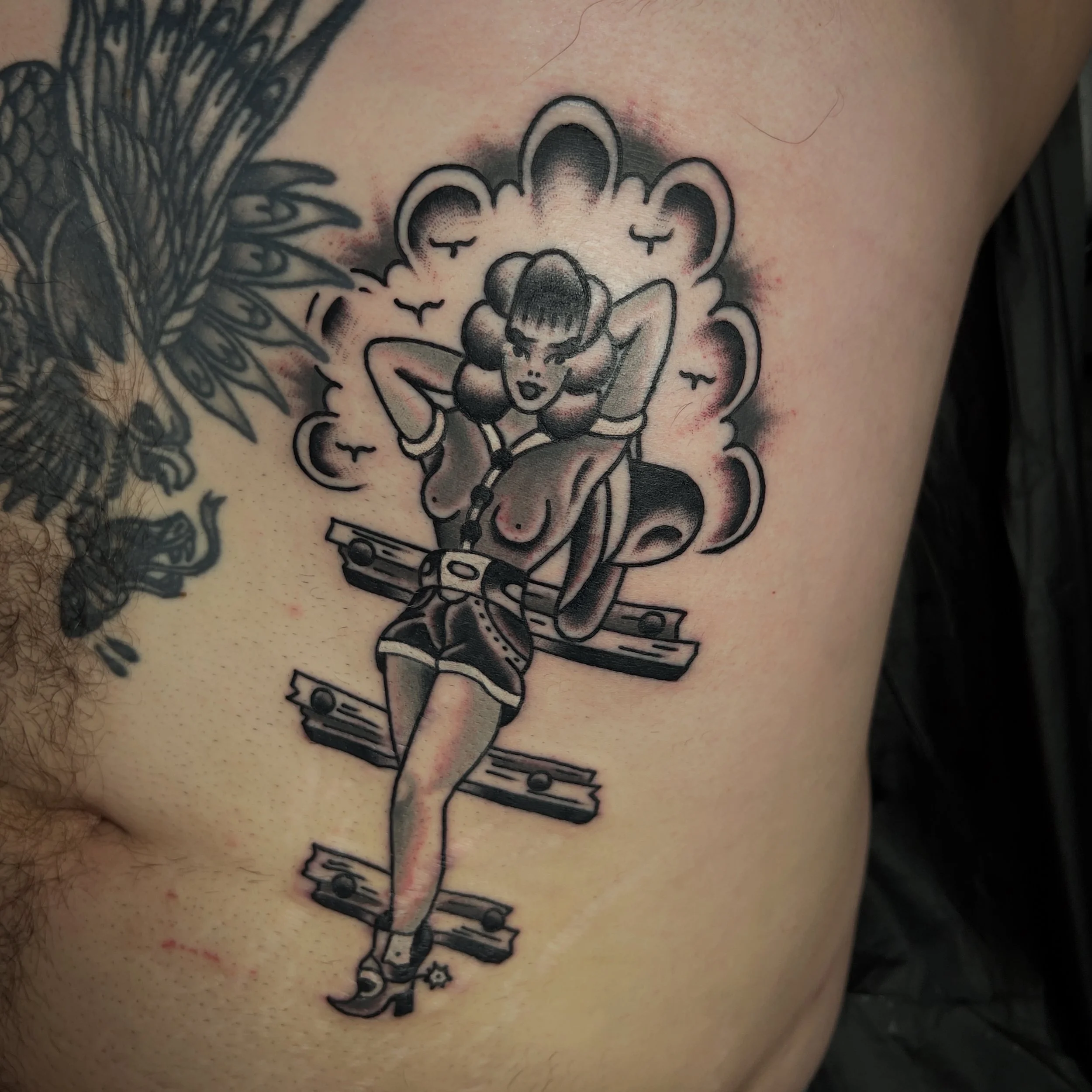 Tattoo of a pin-up girl with pigtails, standing on broken wooden planks, with clouds and birds in the background.
