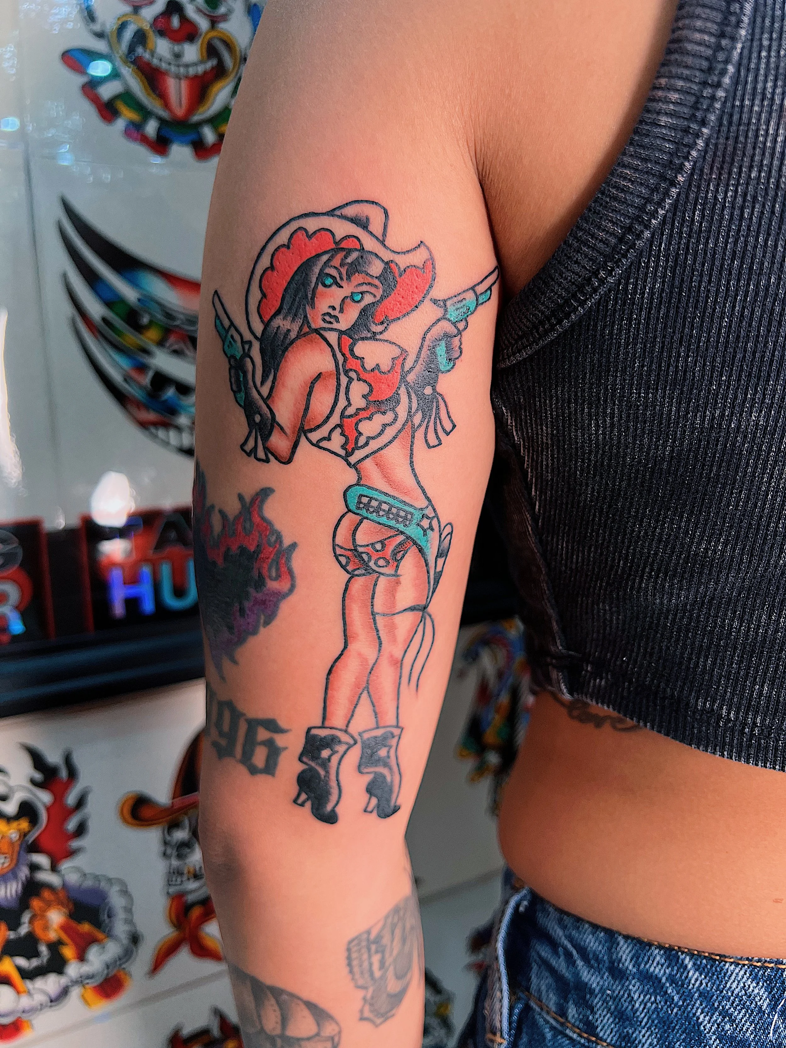 Tattoo of a woman in a cowboy hat holding two guns, wearing a bra, shorts, and cowboy boots, with flames and text on her arm.