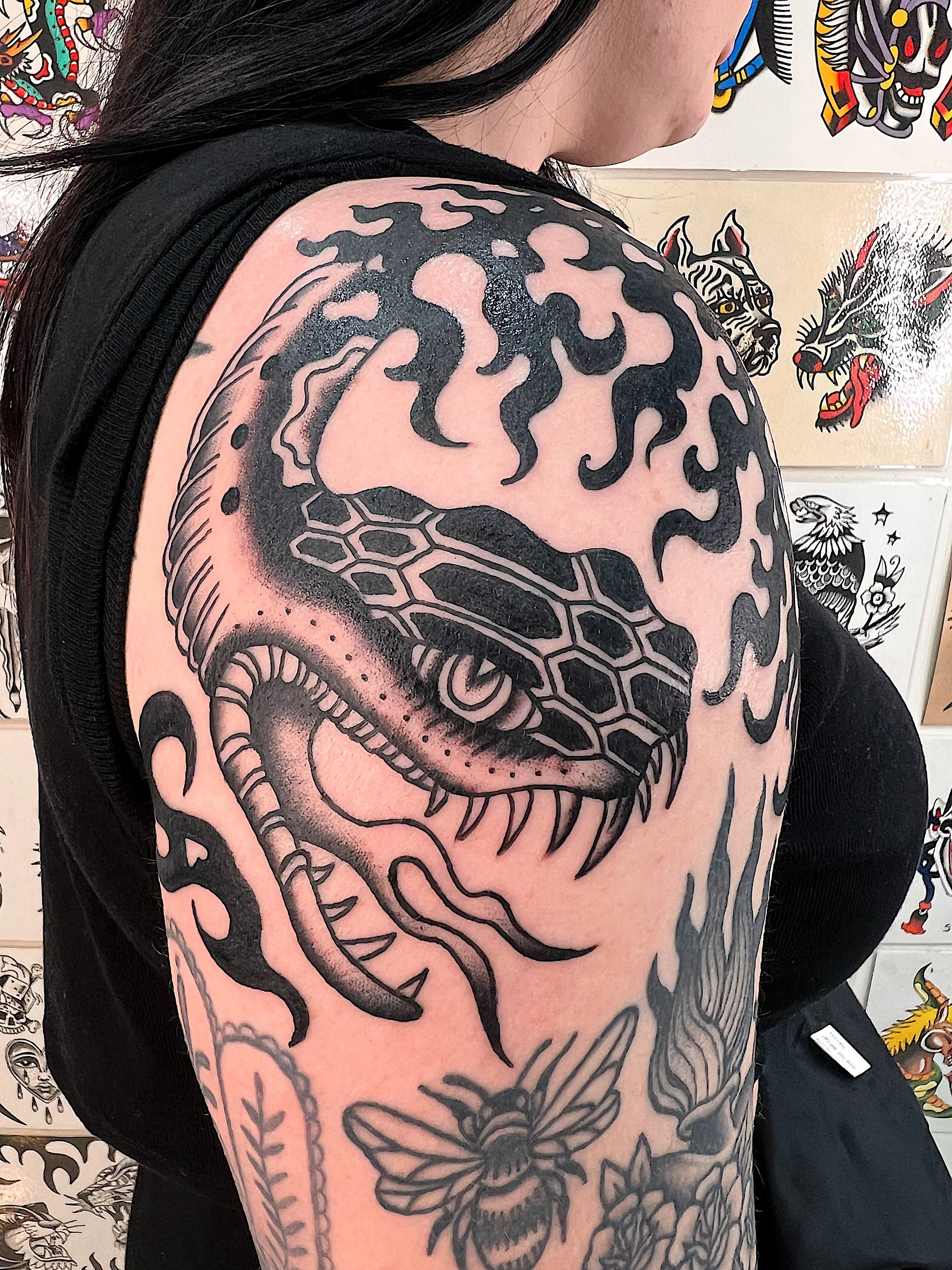Black and gray tattoo of a snake with an open mouth and fangs on a person's shoulder and upper arm, surrounded by floral and insect designs.