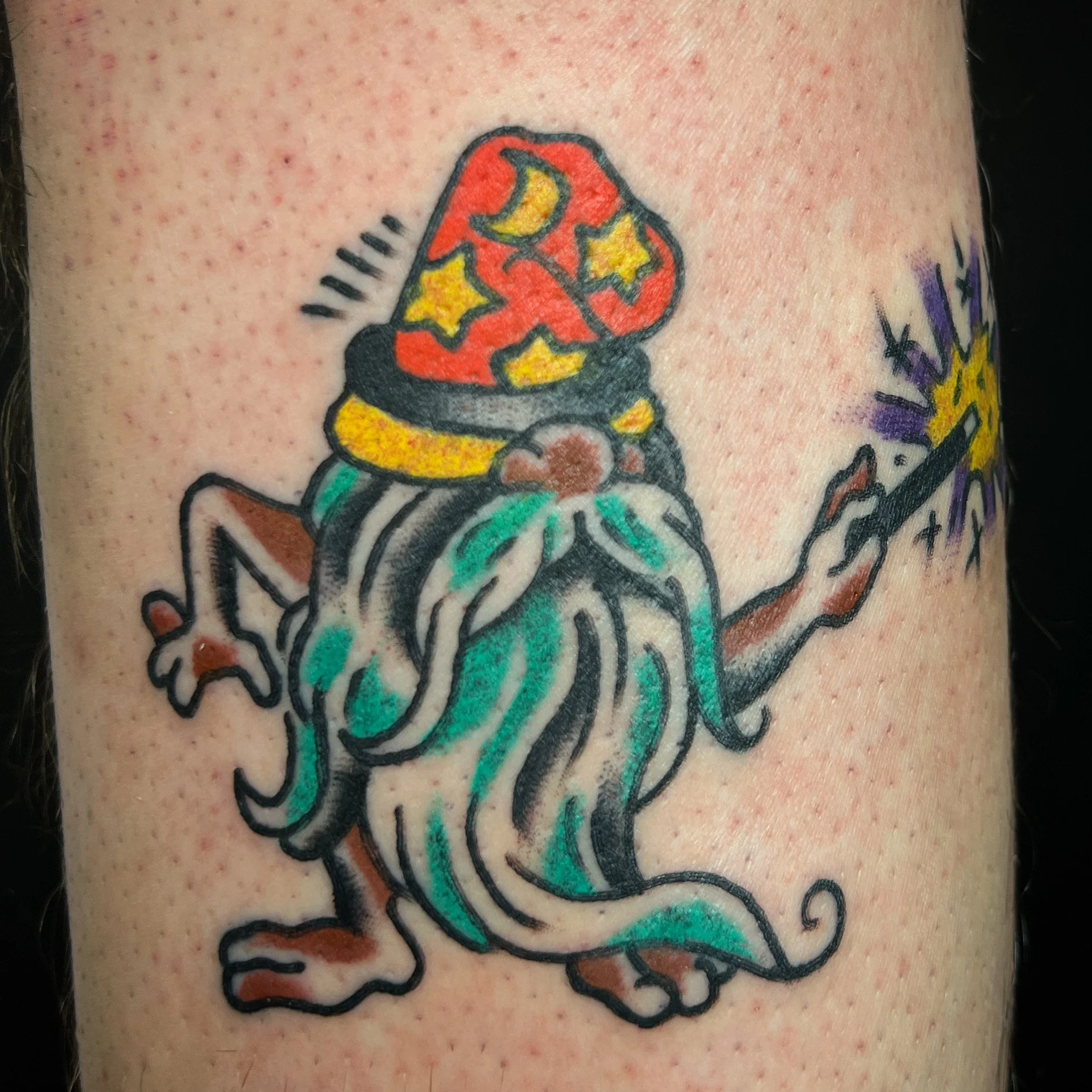 Tattoo of a mythical creature with a wizard hat decorated with stars and a moon, a long flowing beard, and holding a magic wand with sparks.