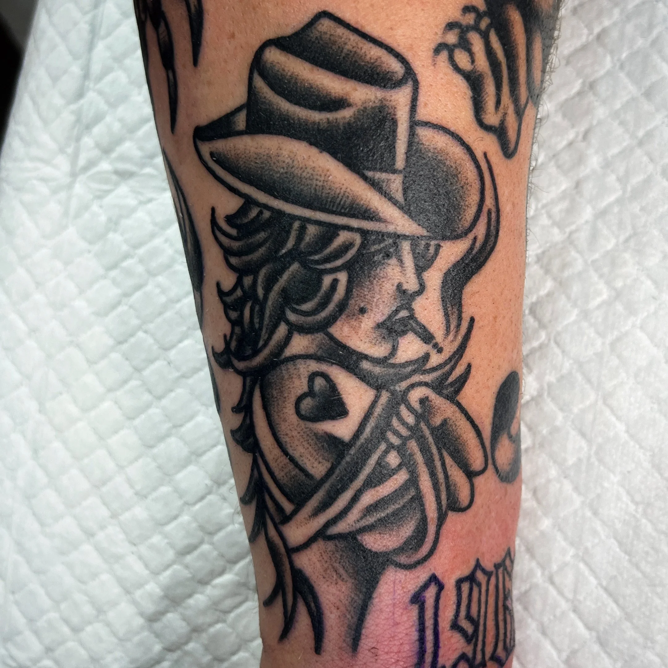 A black and gray tattoo of a woman wearing a wide-brimmed hat, smoking a cigarette, with a heart shape on her chest and the date '1990' tattooed below.