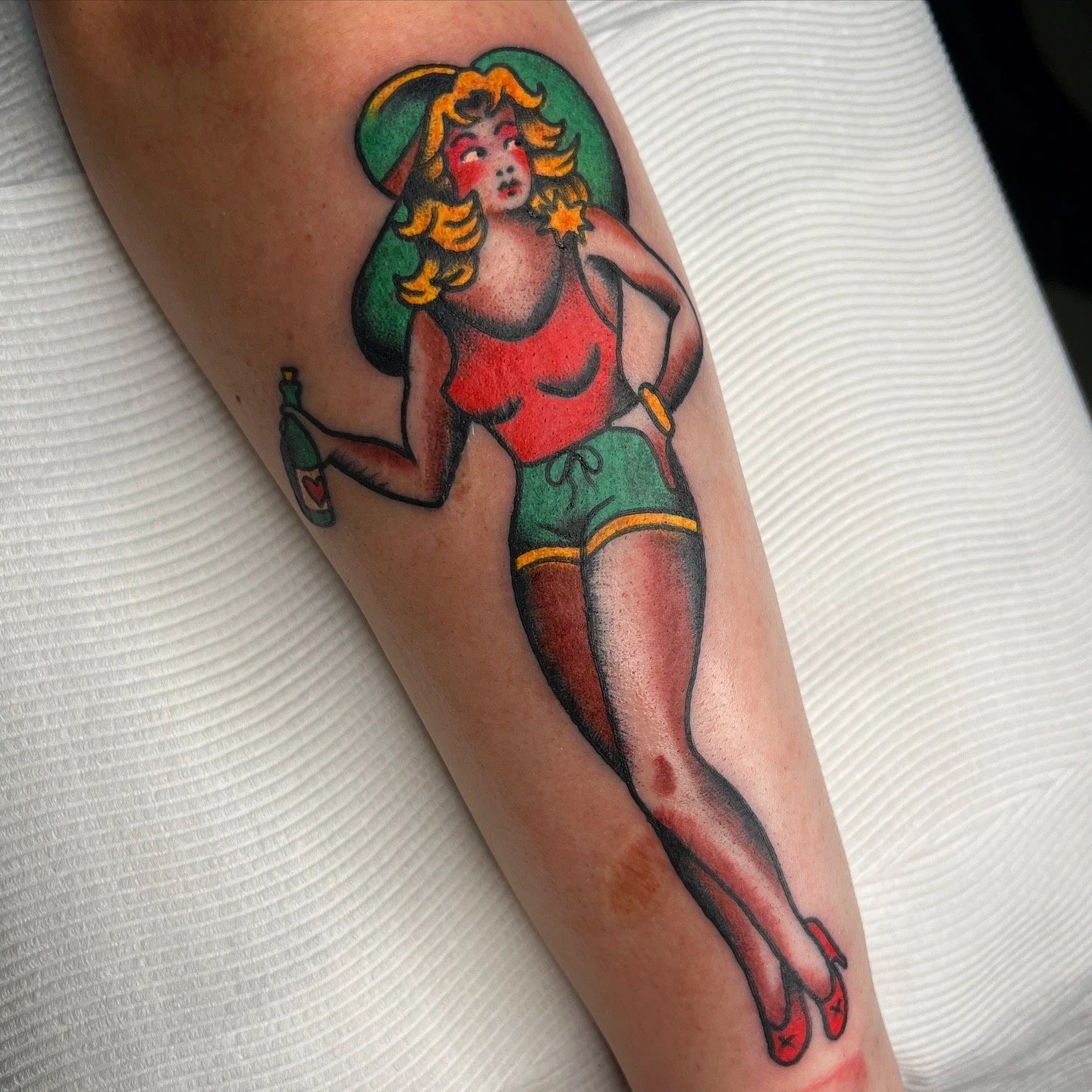 Tattoo of a vintage pin-up style woman with blonde wavy hair, wearing a green wide-brimmed hat, red tank top, green shorts with yellow trim, thigh-high stockings, red high heels, holding a beer bottle, and featuring vibrant colors and bold outlines.