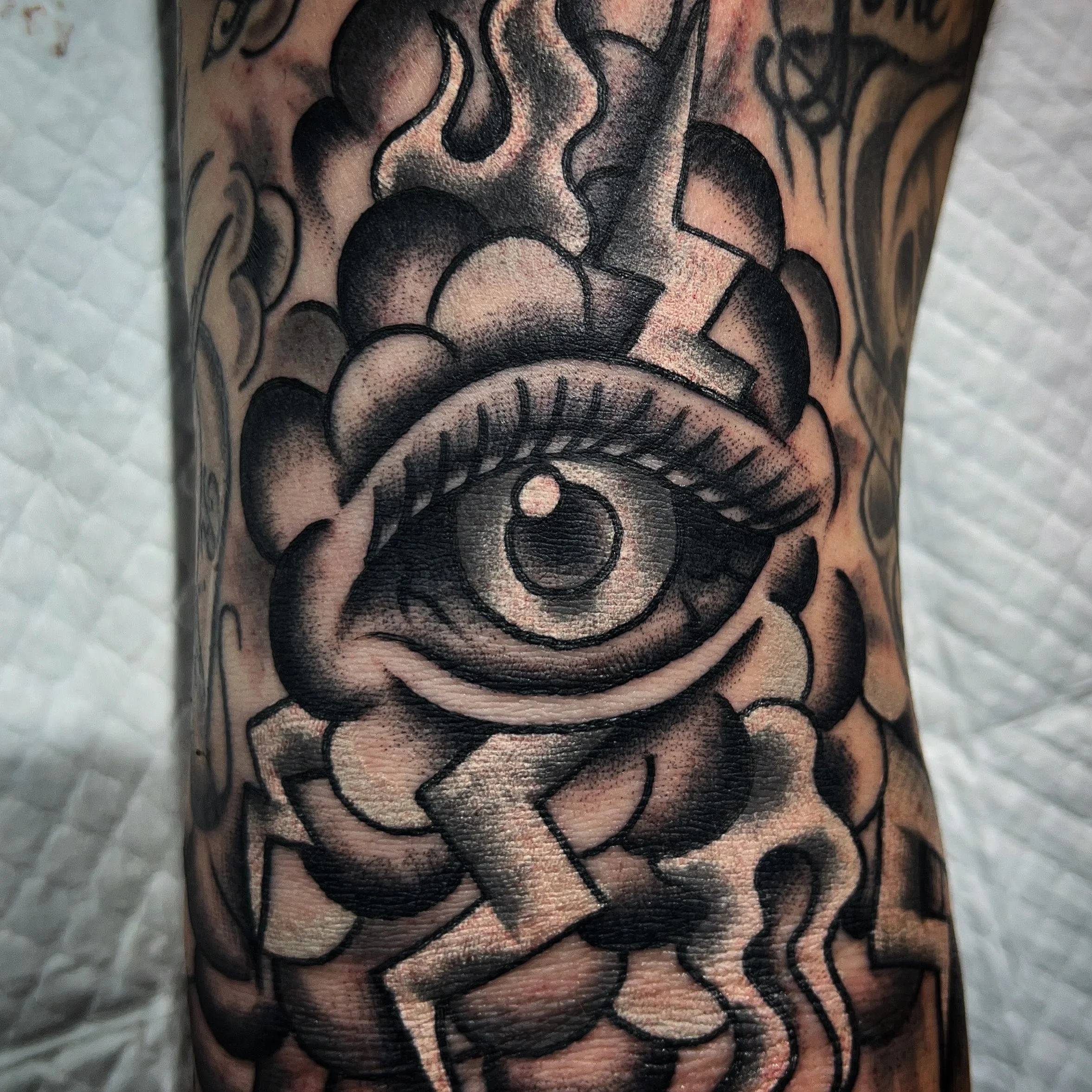 A black and gray tattoo of an eye with eyelashes, surrounded by clouds, with a lightning bolt and a crescent moon within the eye.
