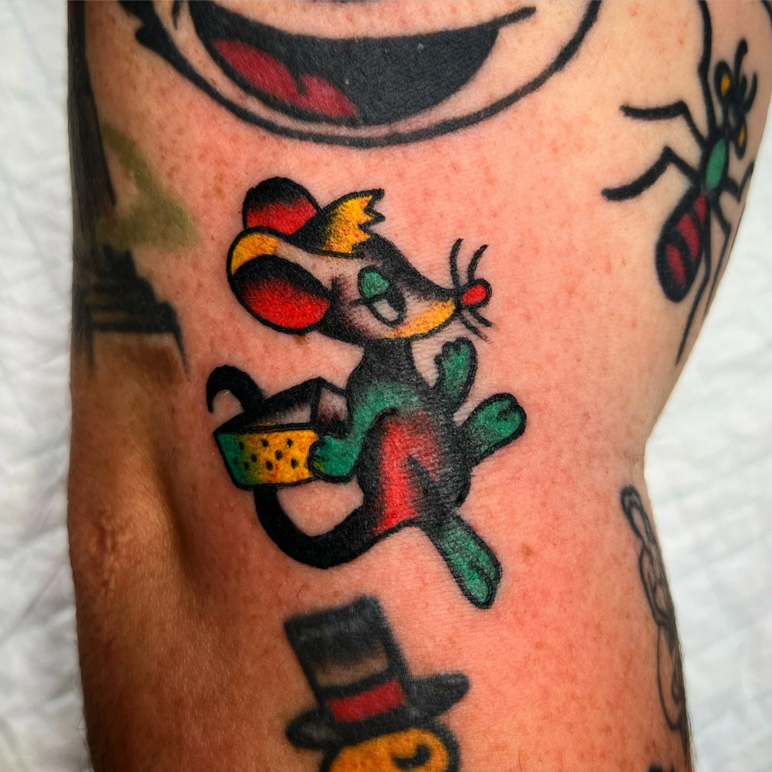 Colorful tattoo of a cartoon mouse with large ears, a yellow patch on one ear, sitting on a cactus, wearing yellow shorts with black dots.