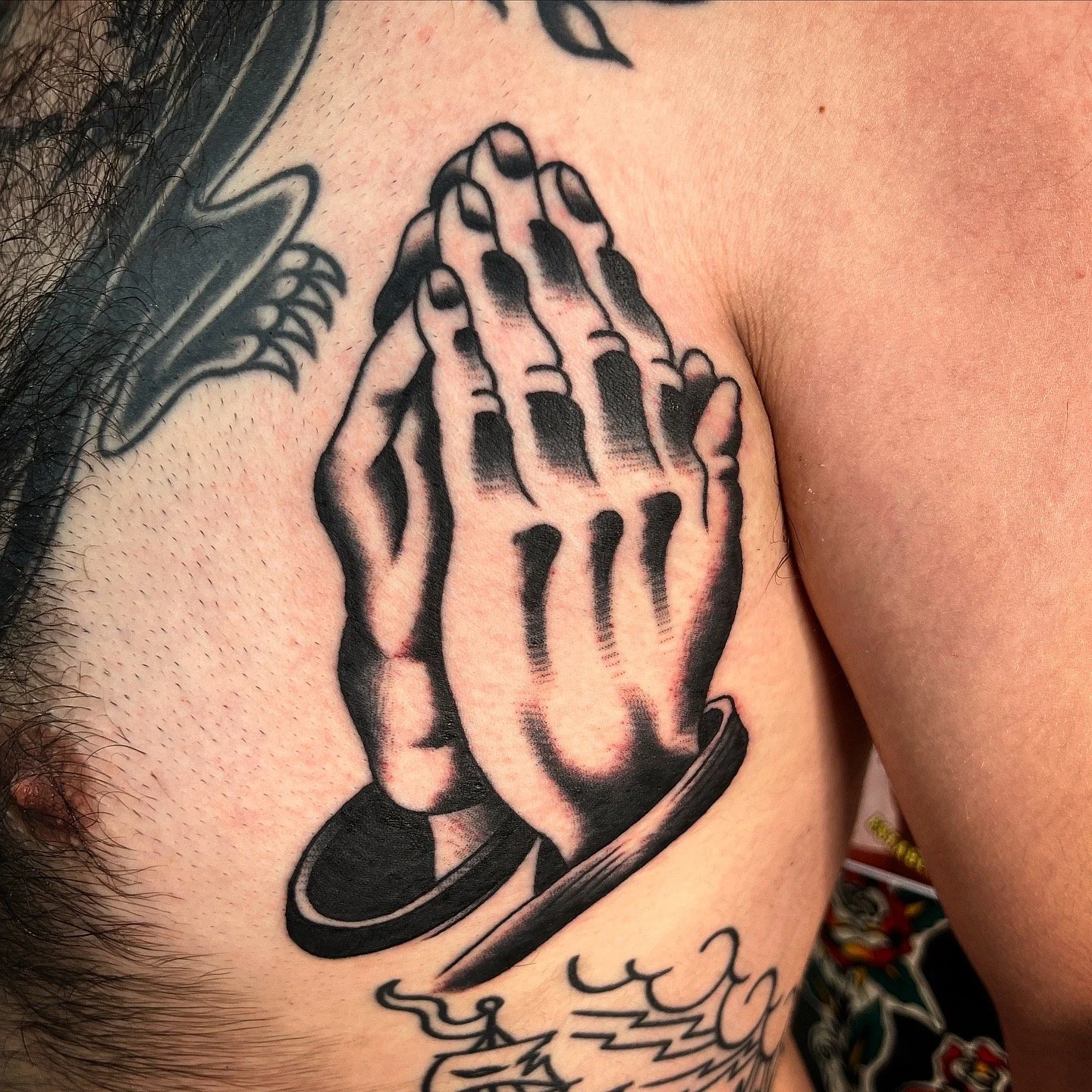 Tattoo of praying hands on a person's chest, with additional tattoo elements visible on the shoulder and chest.