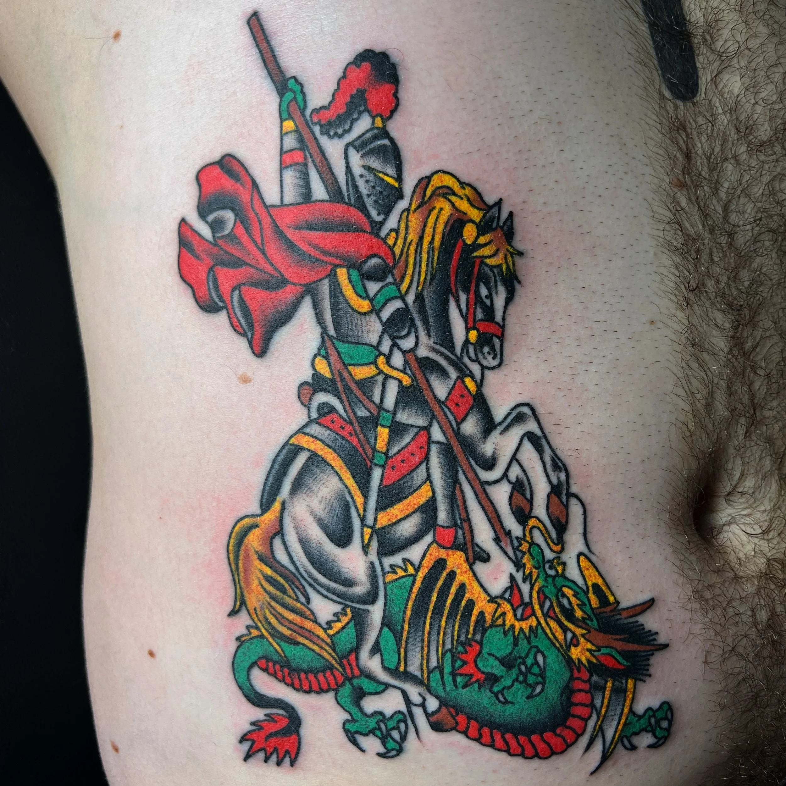 Tattoo of a knight riding a dragon, colored in black, red, yellow, and green, with armor, a shield, and a helmet with a plume.