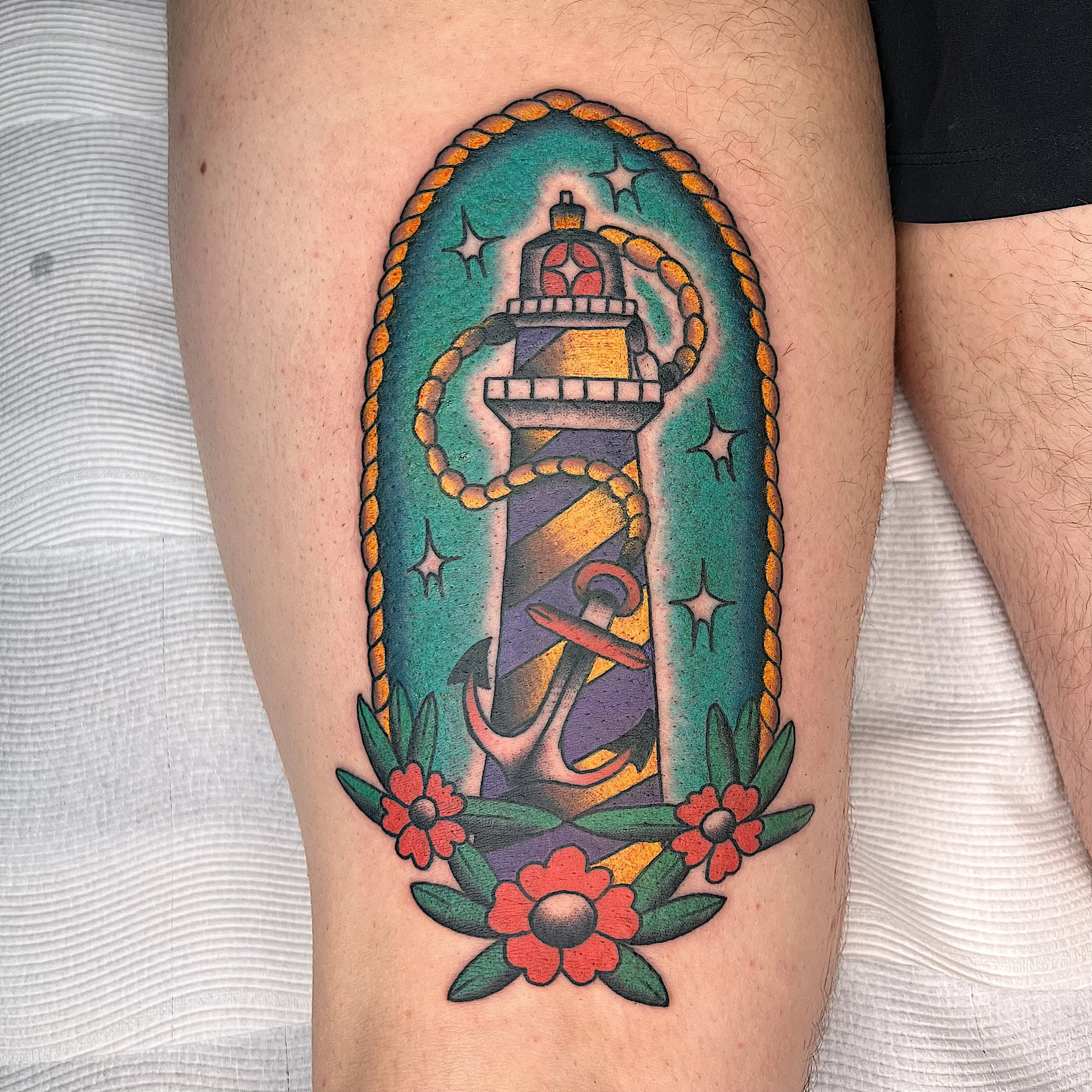 Tattoo of a lighthouse with an anchor, flowers, and a background of stars.