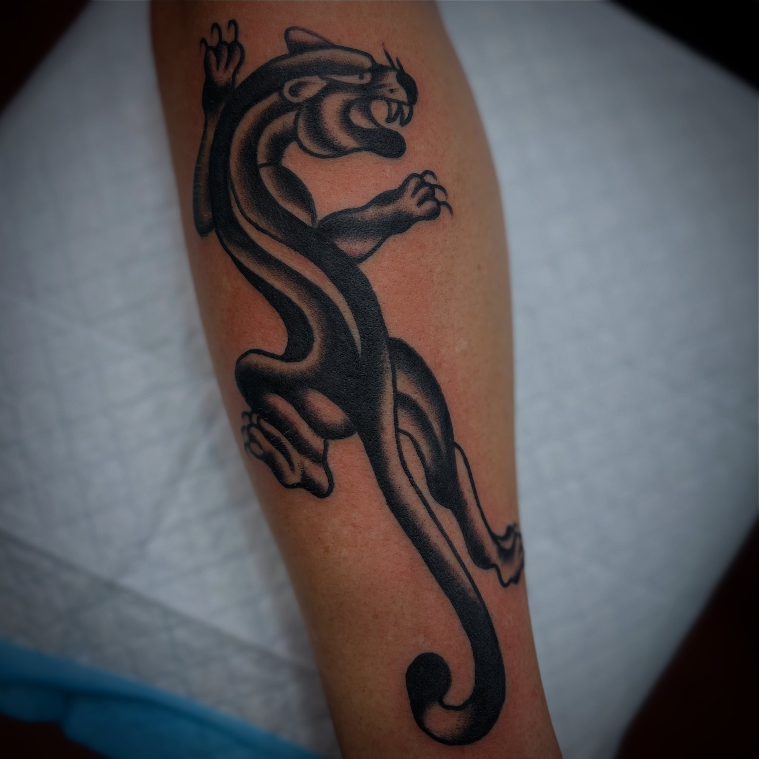 Black and gray tattoo of a fierce, roaring tiger with an elongated tail on a person's upper arm.