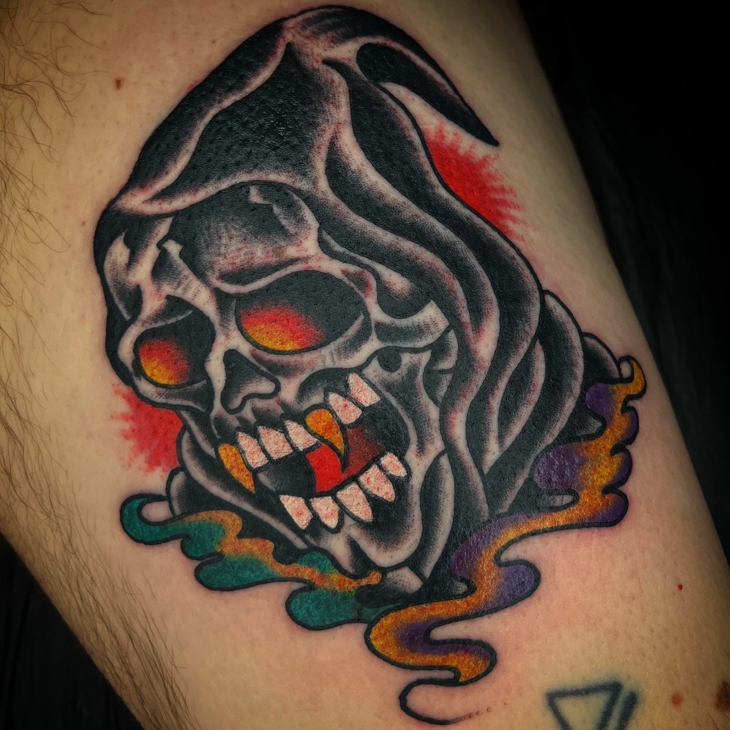 A colorful tattoo of a skull with a hooded figure, with fiery eyes and mouth, surrounded by swirling smoke in green, purple, orange, and yellow.