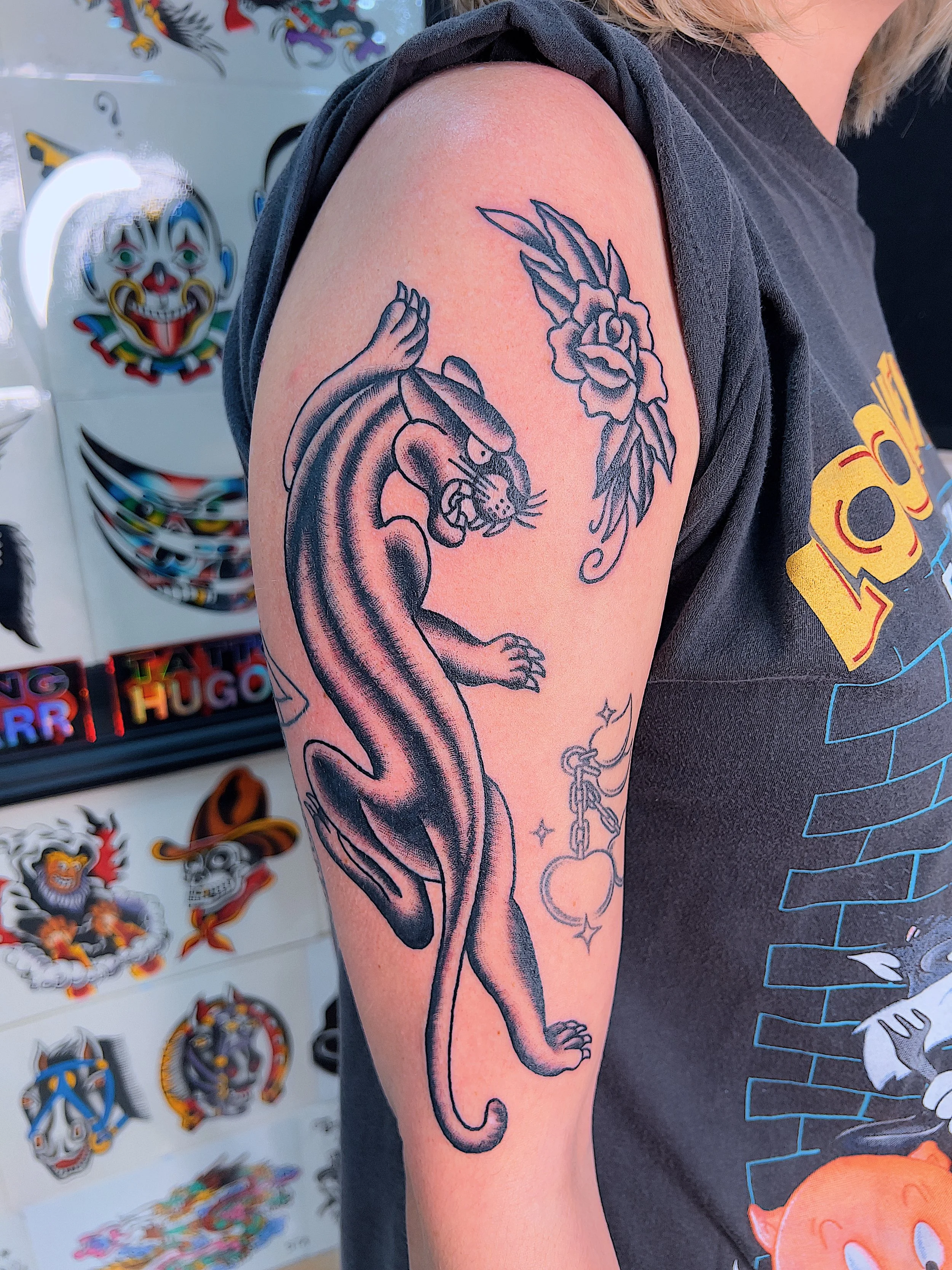 Tattoo of a fierce panther with claws extended on upper arm, next to a flower and a chain with a lock and star charms, on a person's upper arm.