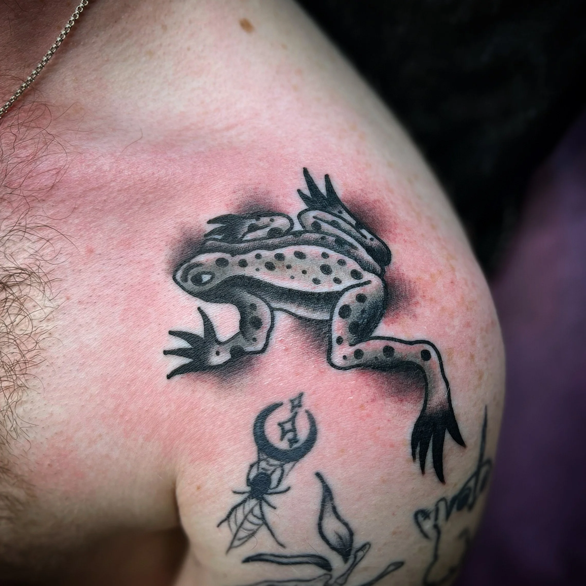 A black and gray tattoo of a frog on a person's upper arm, with additional small decorative designs and text.