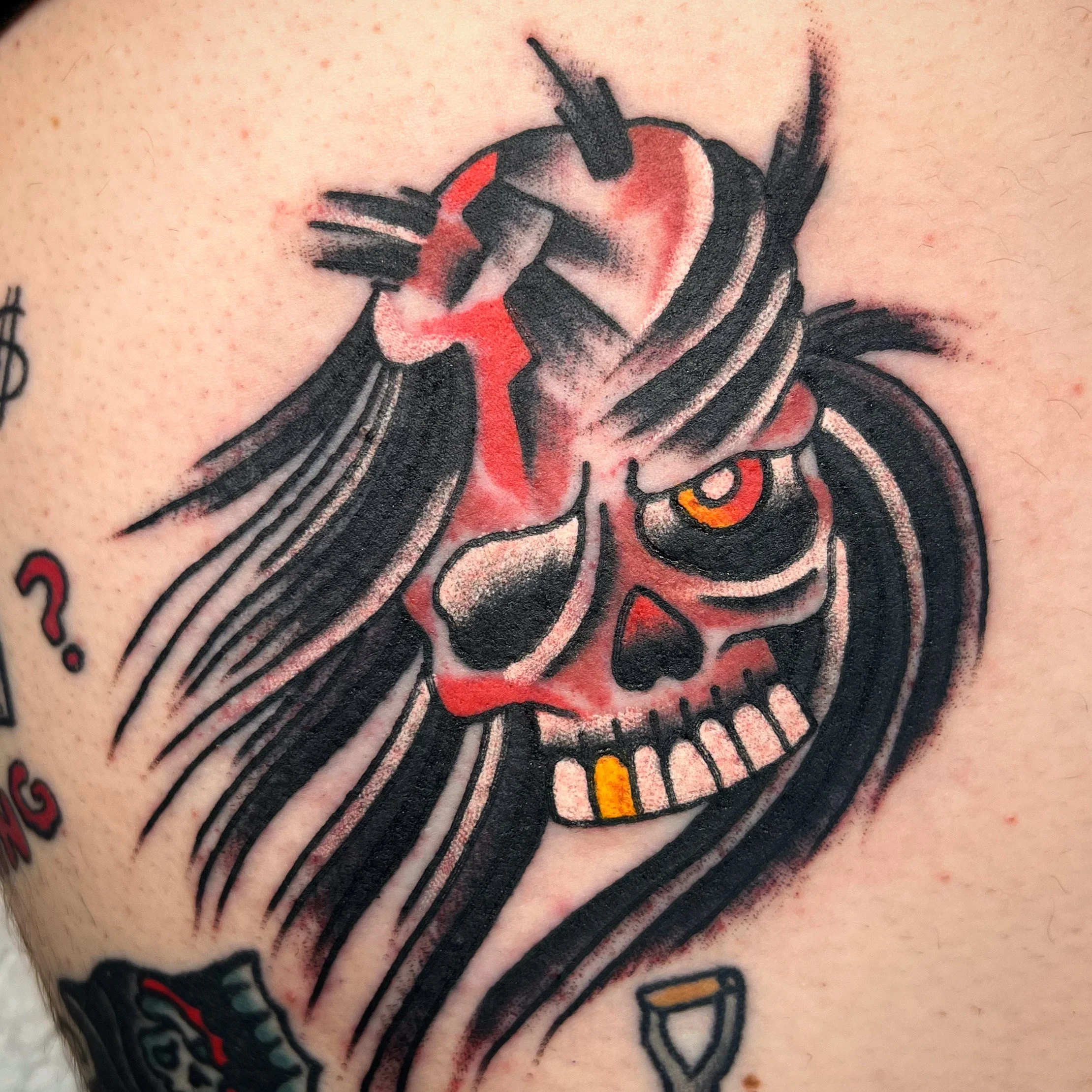 Tattoo of a skeleton with long black hair, red and black shading, an eye with a yellow iris, and a cigar in its mouth.