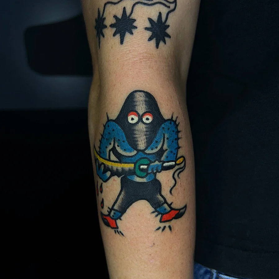 A colorful tattoo of a cartoon rat wearing black clothing, red shoes, and holding a sword. The rat has white eyes with red around them and is standing in a confident pose.