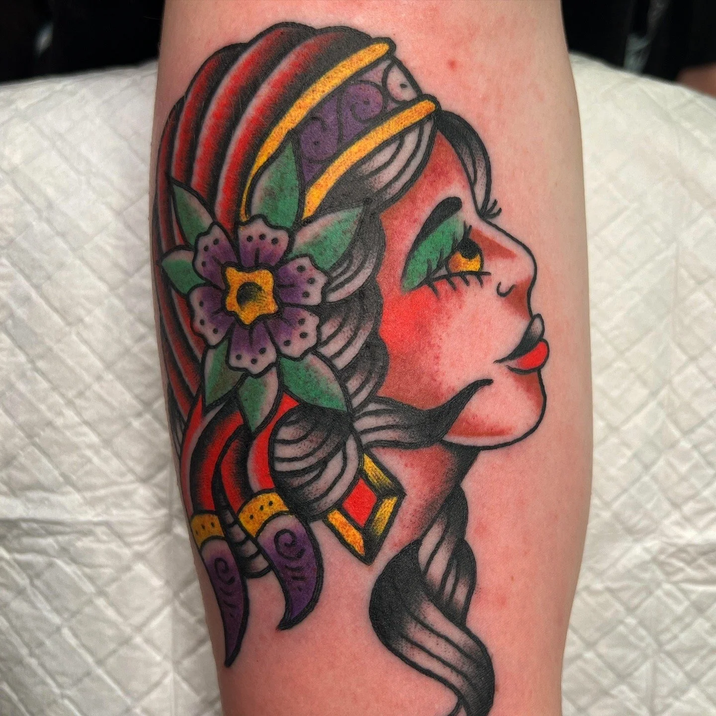 Tattoo of a woman's face in profile with long flowing hair, colorful flowers, leaves, and jewelry, featuring bold outlines and vibrant colors.