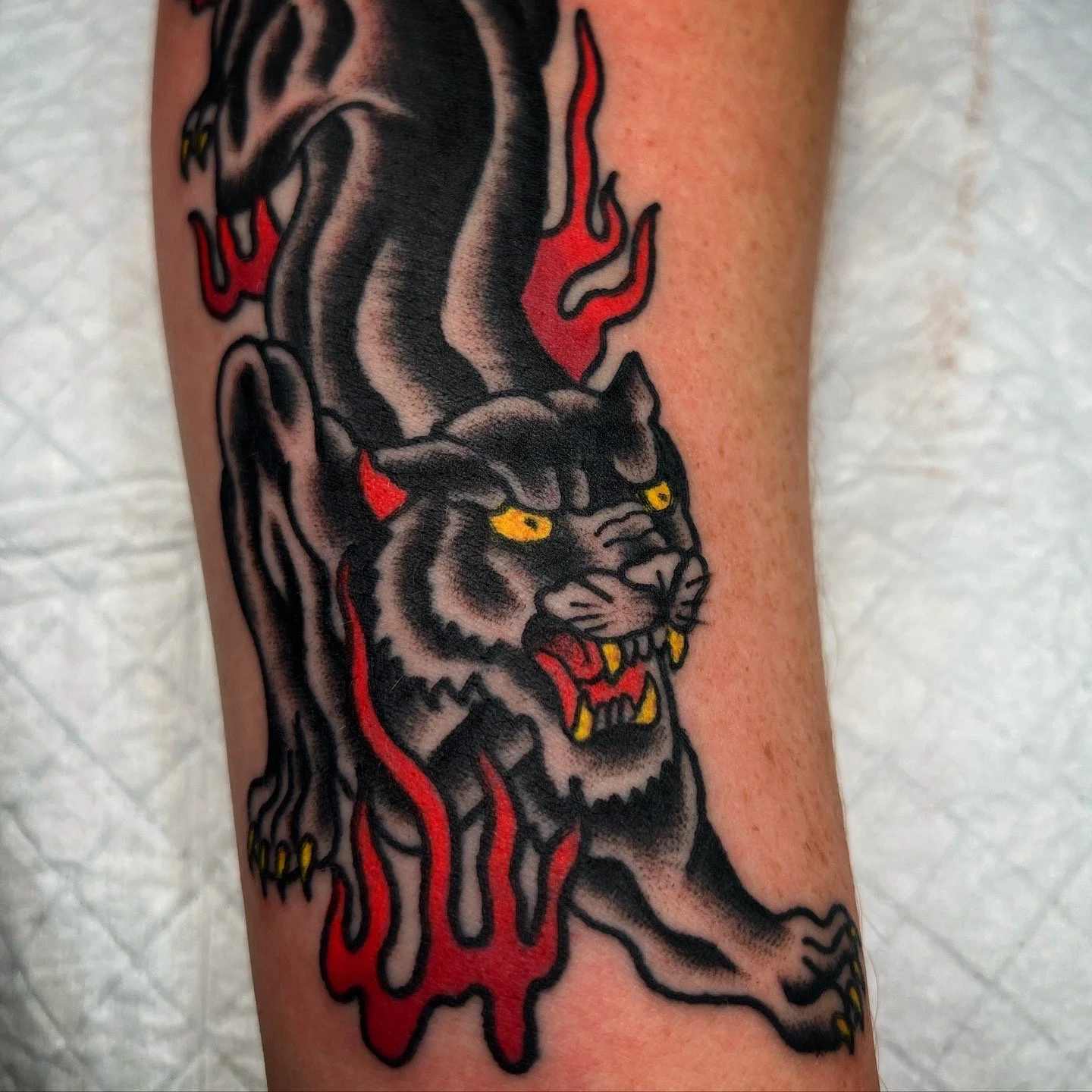 Tattoo of a fierce black panther with yellow eyes, showing teeth and claws, with red flames around its neck and red details throughout the body.