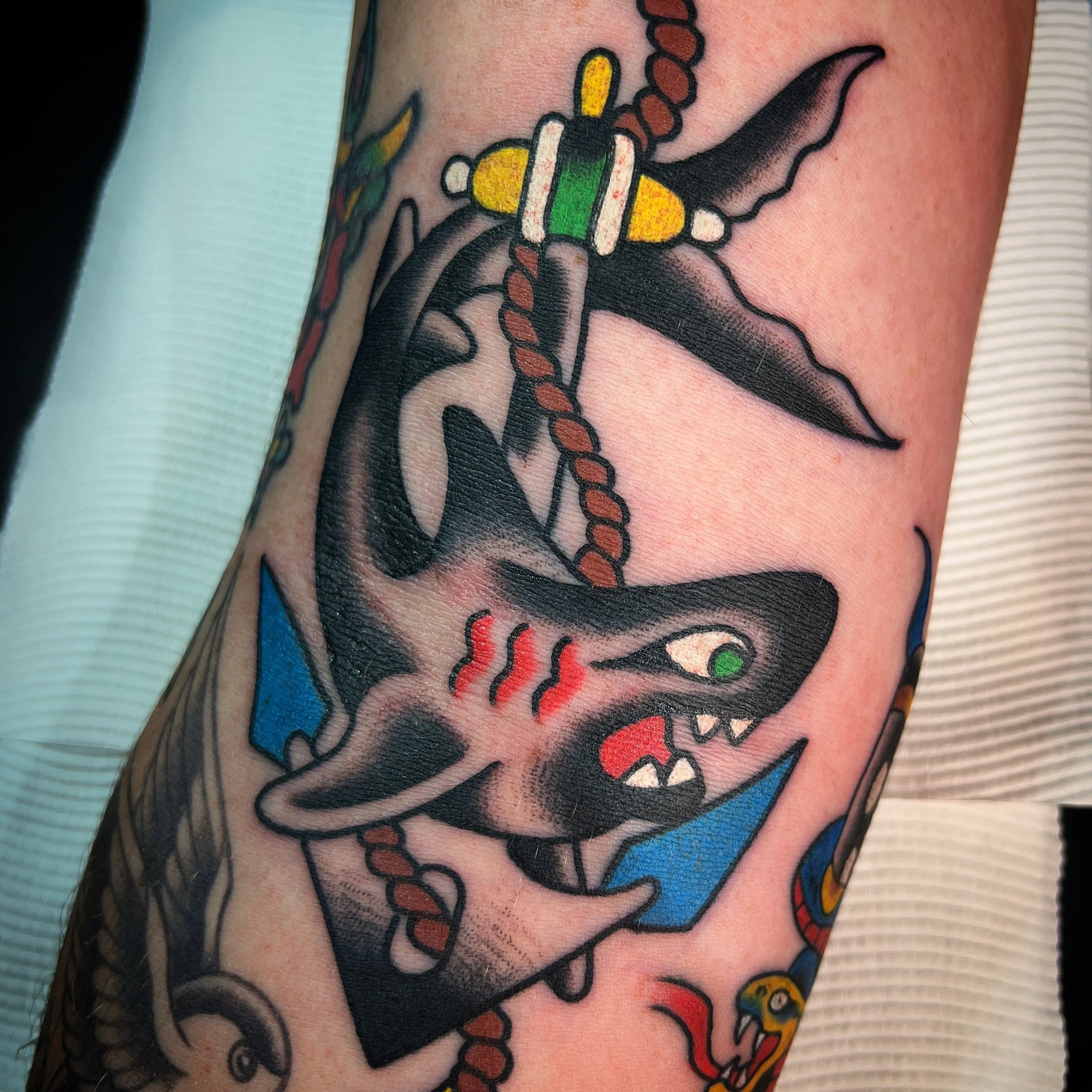Tattoo of a black cat with green eyes, open mouth showing teeth, red steam lines on chest, inside a blue triangle, with a traditional style beaded necklace and a dagger piercing through the triangle.