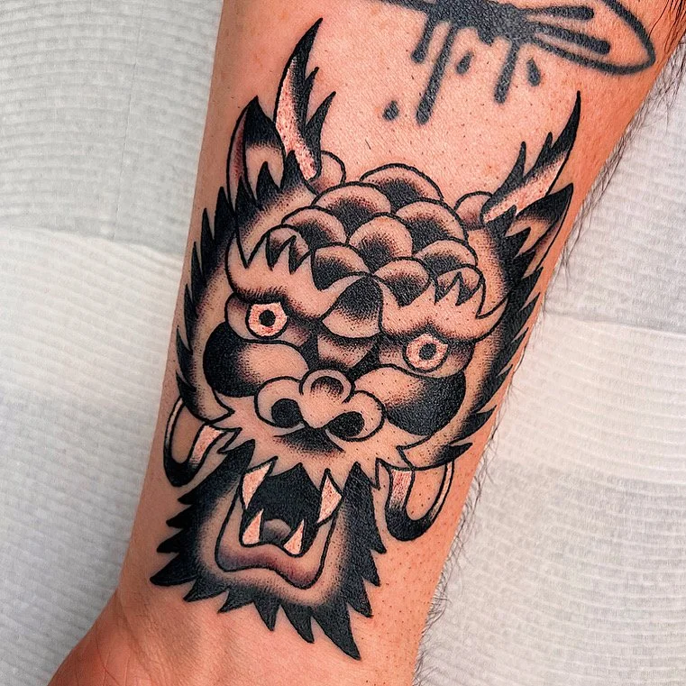 Black and gray tattoo of a fierce dragon's head with detailed scales, sharp teeth, and piercing eyes on a person's arm.