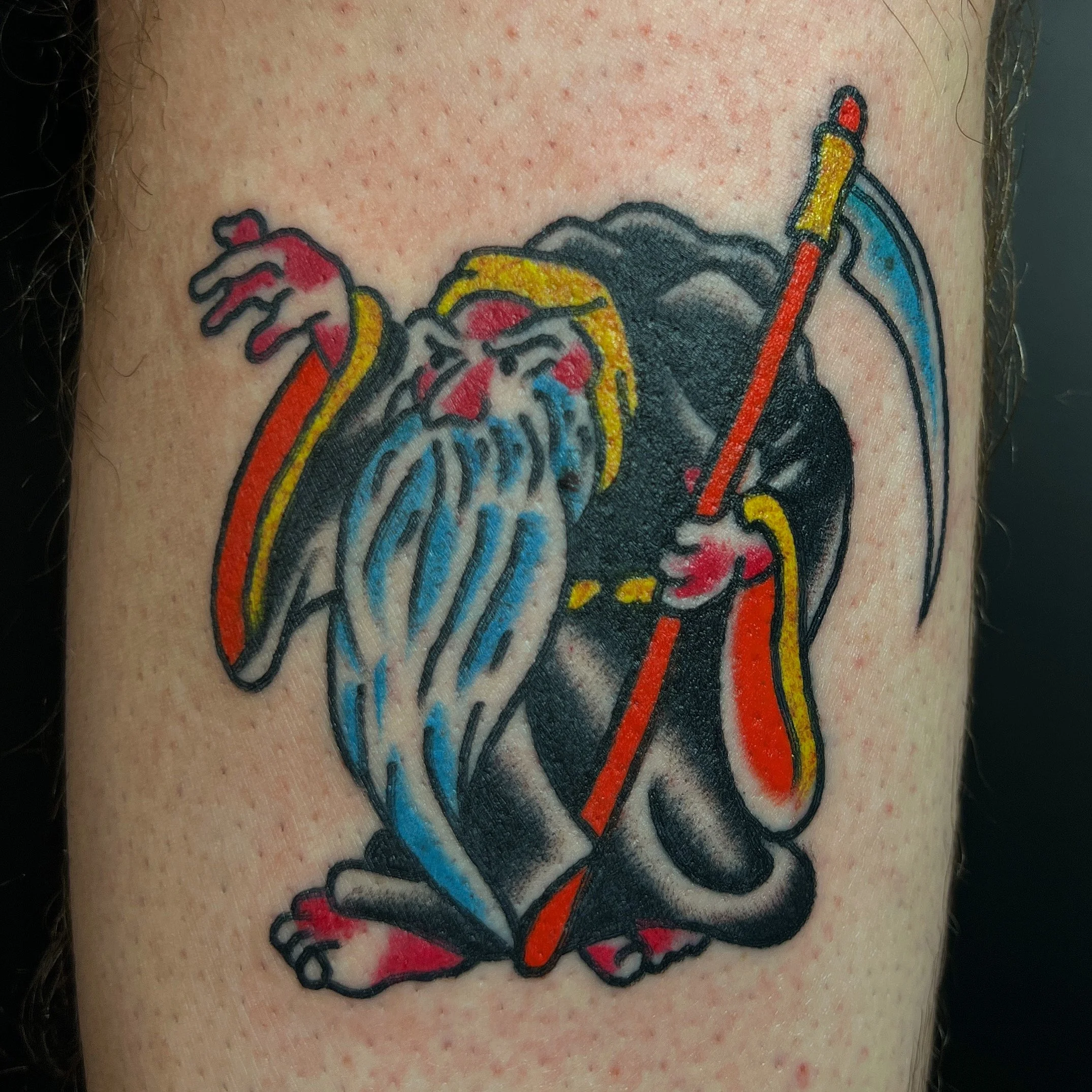 Colorful tattoo of a grim reaper with a scythe, dressed in black with yellow and red accents, holding a blue and white shield, on skin.