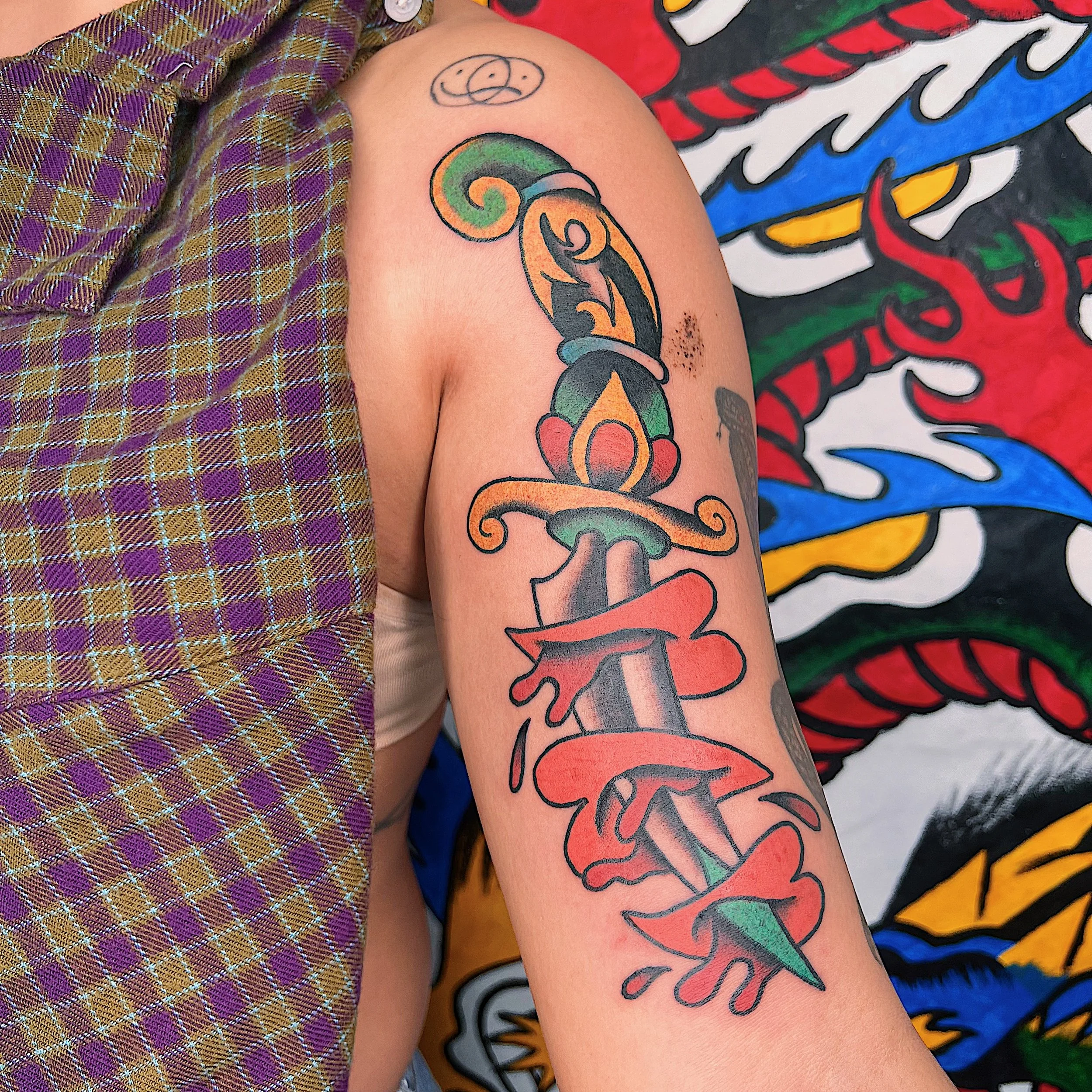 Close-up of a person's arm with a colorful tattoo of a sword wrapped with red ribbon, decorated with teardrops, against a background of a painted wall with bold colors and patterns.