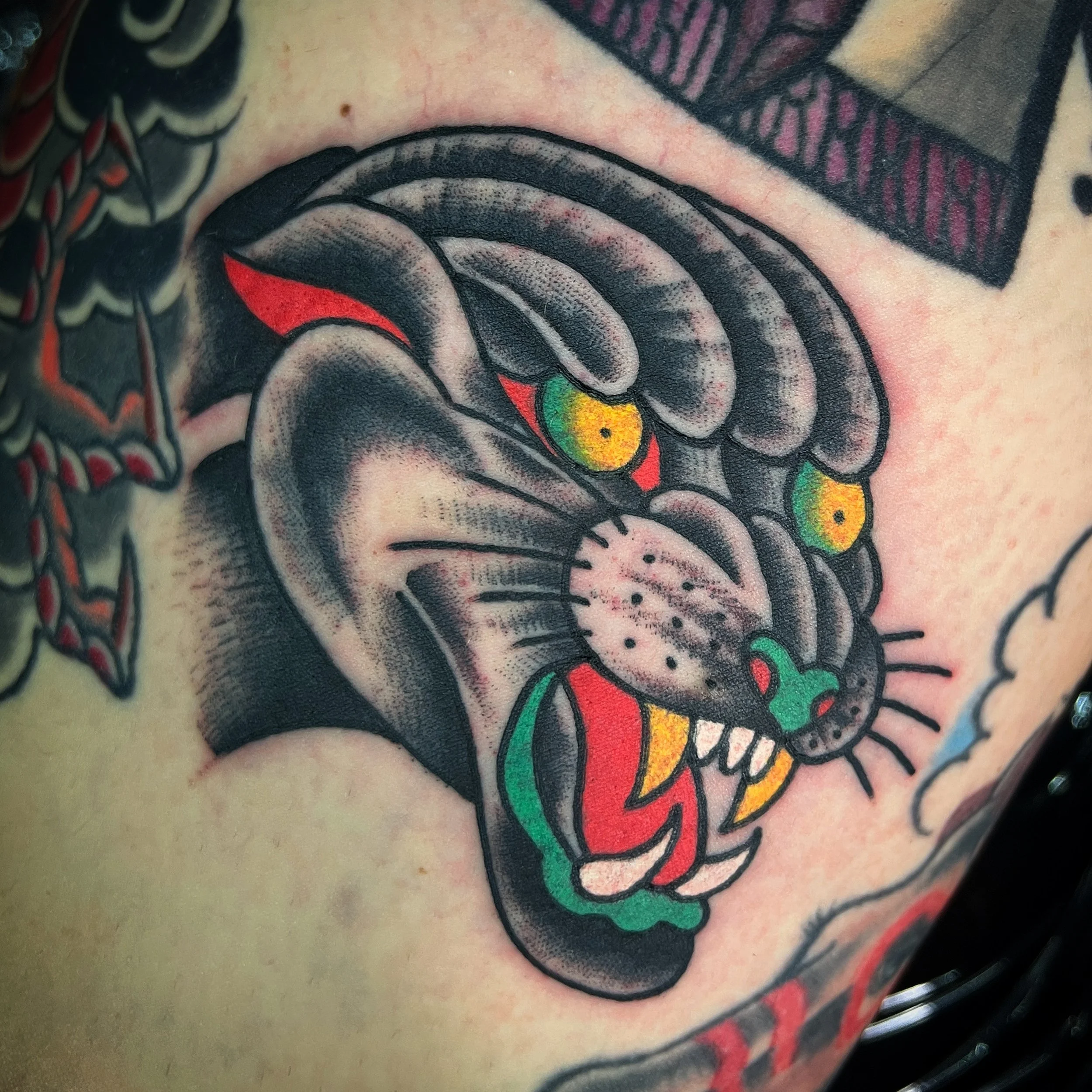 Colorful traditional tattoo of a fierce lion's face with yellow eyes, green nose, red tongue, and black, gray, and red details on skin.