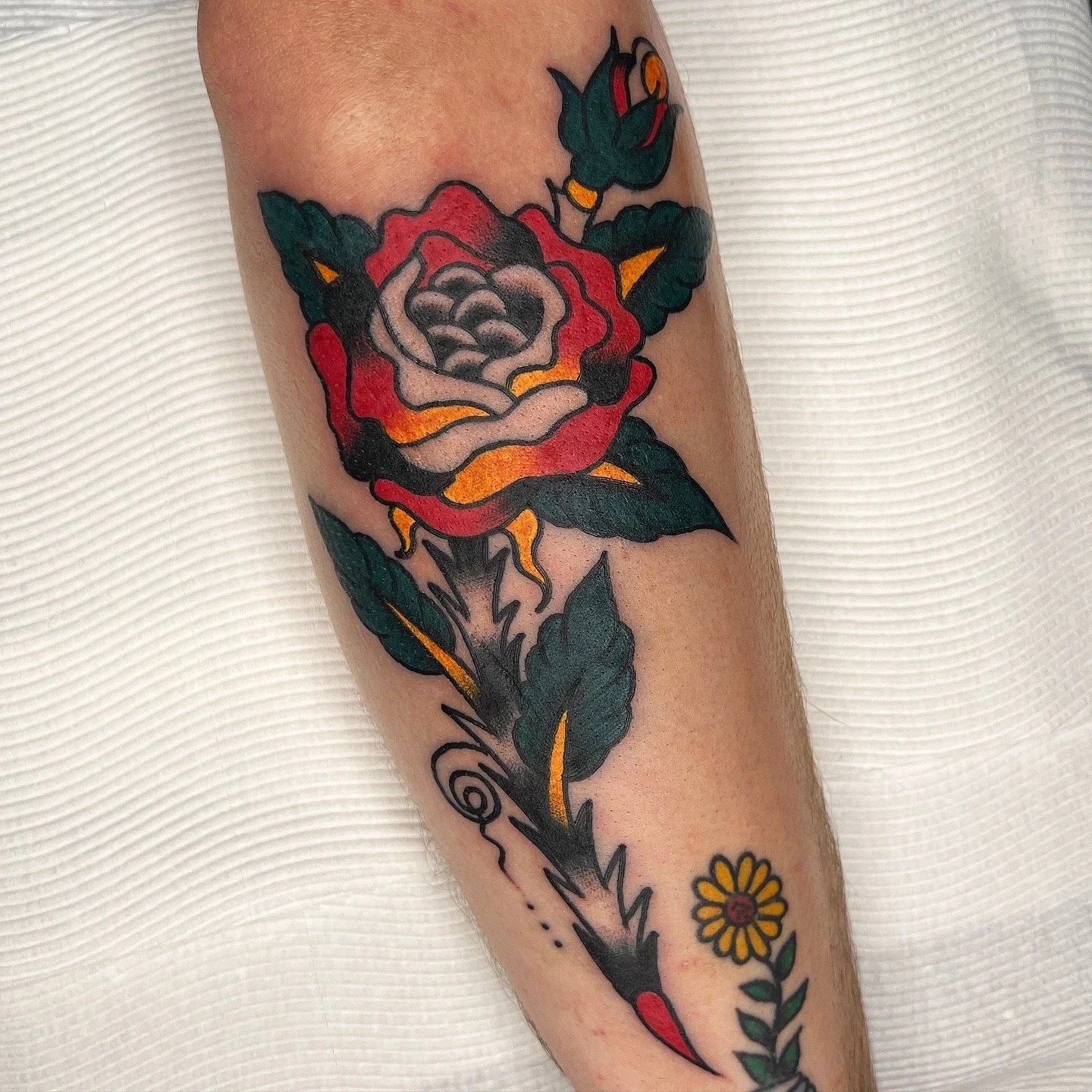 A colorful traditional tattoo of a large red, pink, and orange rose with green leaves on a person's forearm, with a smaller yellow flower below.