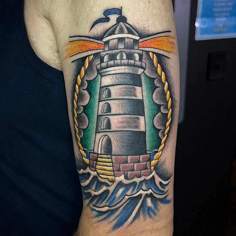 Tattoo of a lighthouse with waves at its base, surrounded by a rope border, and a sunset or sunrise sky in the background.