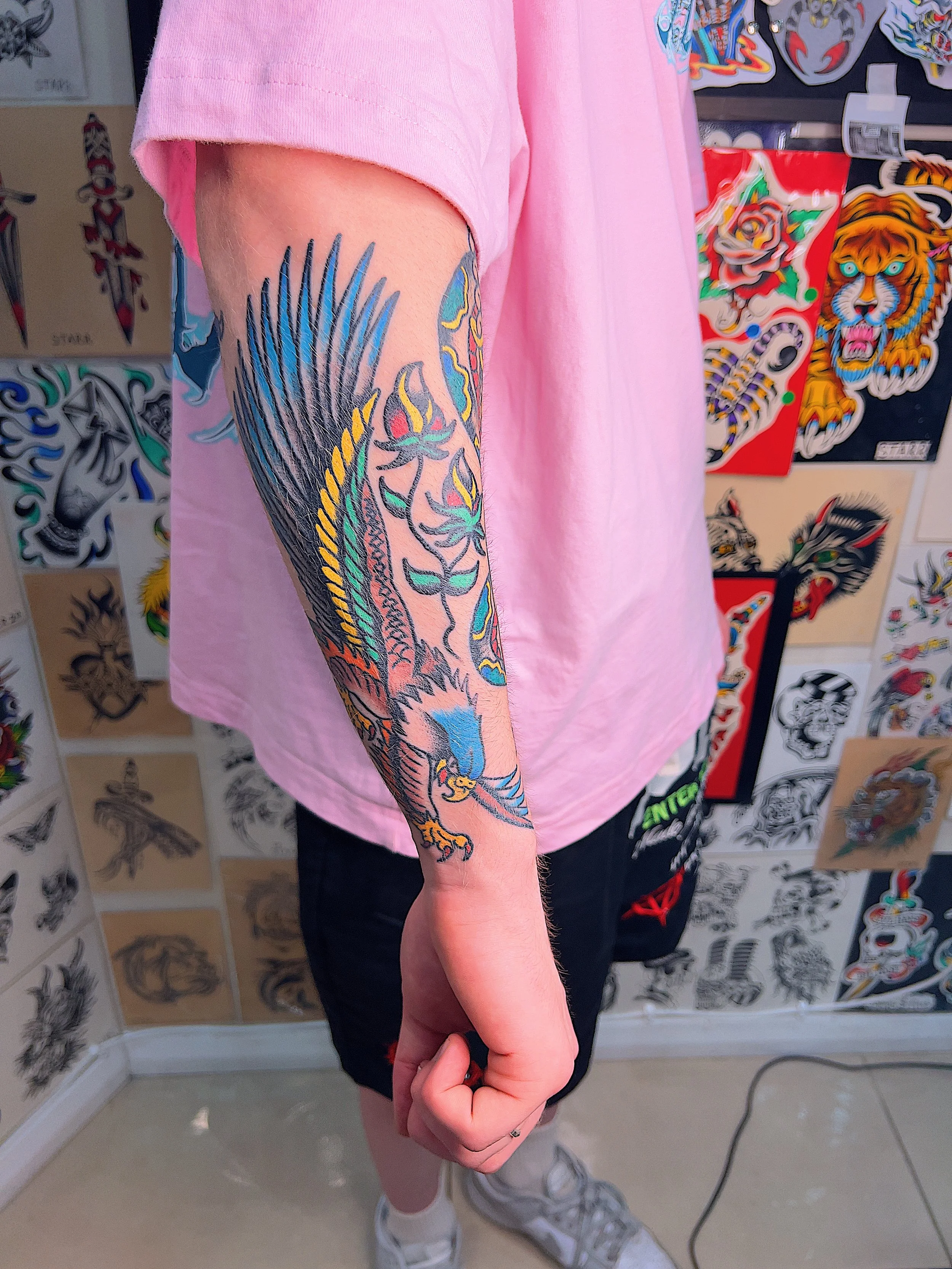 Person with a colorful tattoo sleeve of a bird, flames, and other designs on their left arm, standing in front of tattoo flash art on the wall.
